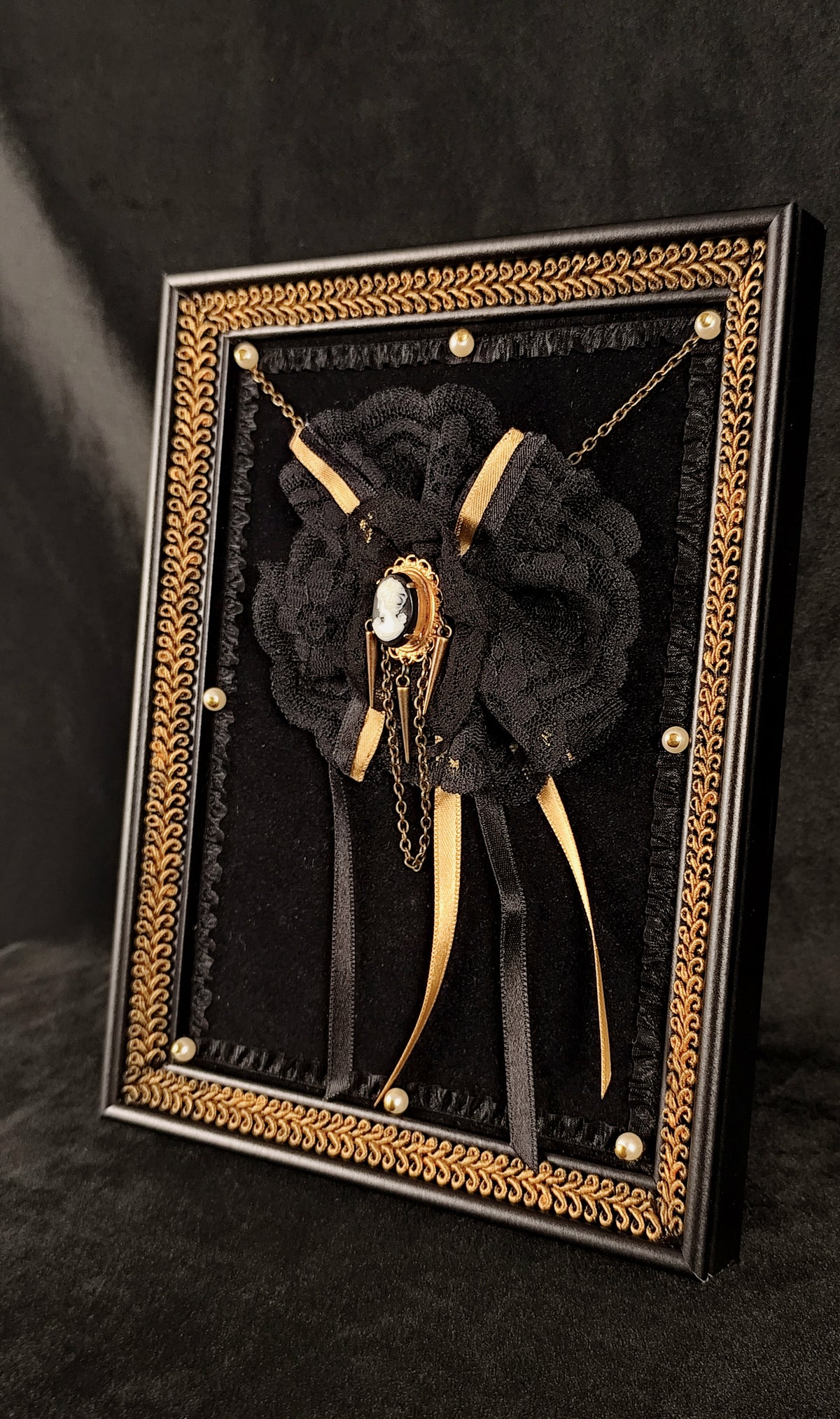 A gothic-style black lace rose brooch with gold chains, ribbons, and a central white stone, presented in an ornate black and gold rectangular display frame.