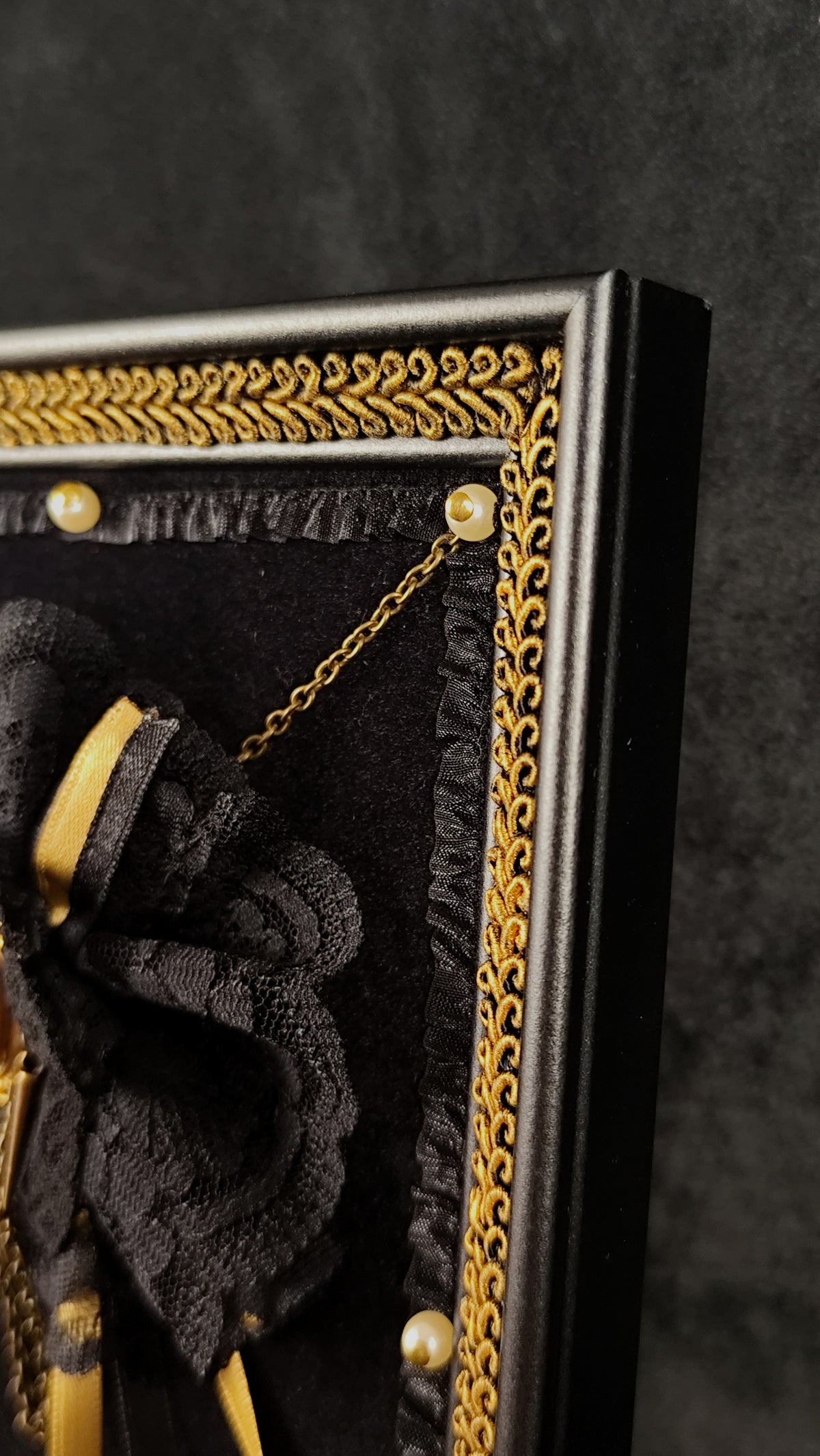Close-up of a black decorative wall art piece with an ornate gold border, black fabric rosette, and gold chain details against a dark textured background.