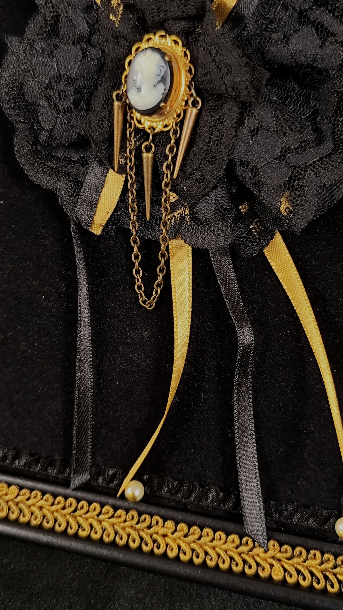 A close-up of a black lace and velvet brooch with gold-toned chains, a central oval cameo-style face detail, and ribbon accents on a black background.