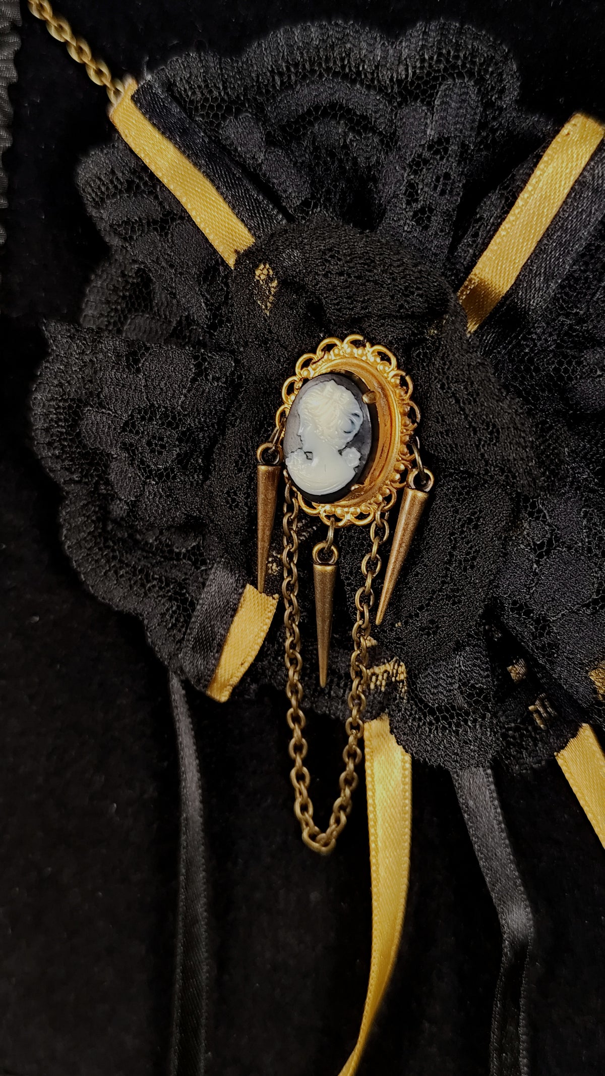 A close-up of a black lace and satin floral accessory with gold trim, ribbon accents, and a central oval cameo brooch on a black background.