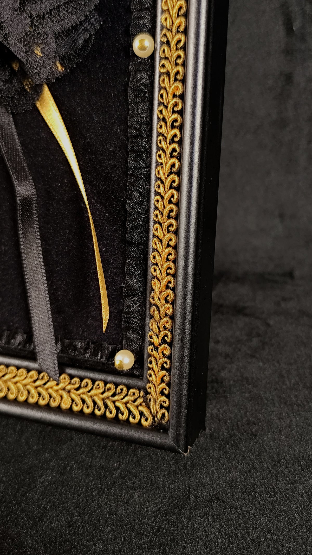 Close-up of a black decorative framed wall piece with ornate gold scrollwork trim, black fabric, ribbon, and pearl-like accents on a dark textured background.
