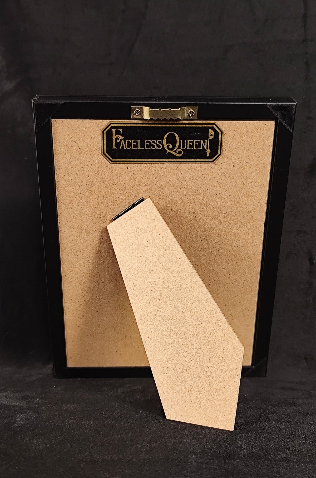 A black-framed picture frame with a tan cardboard backing and a matching leaning insert, photographed against a dark textured background. The brand name 'FACELESS QUEEN' is printed on the top center label.