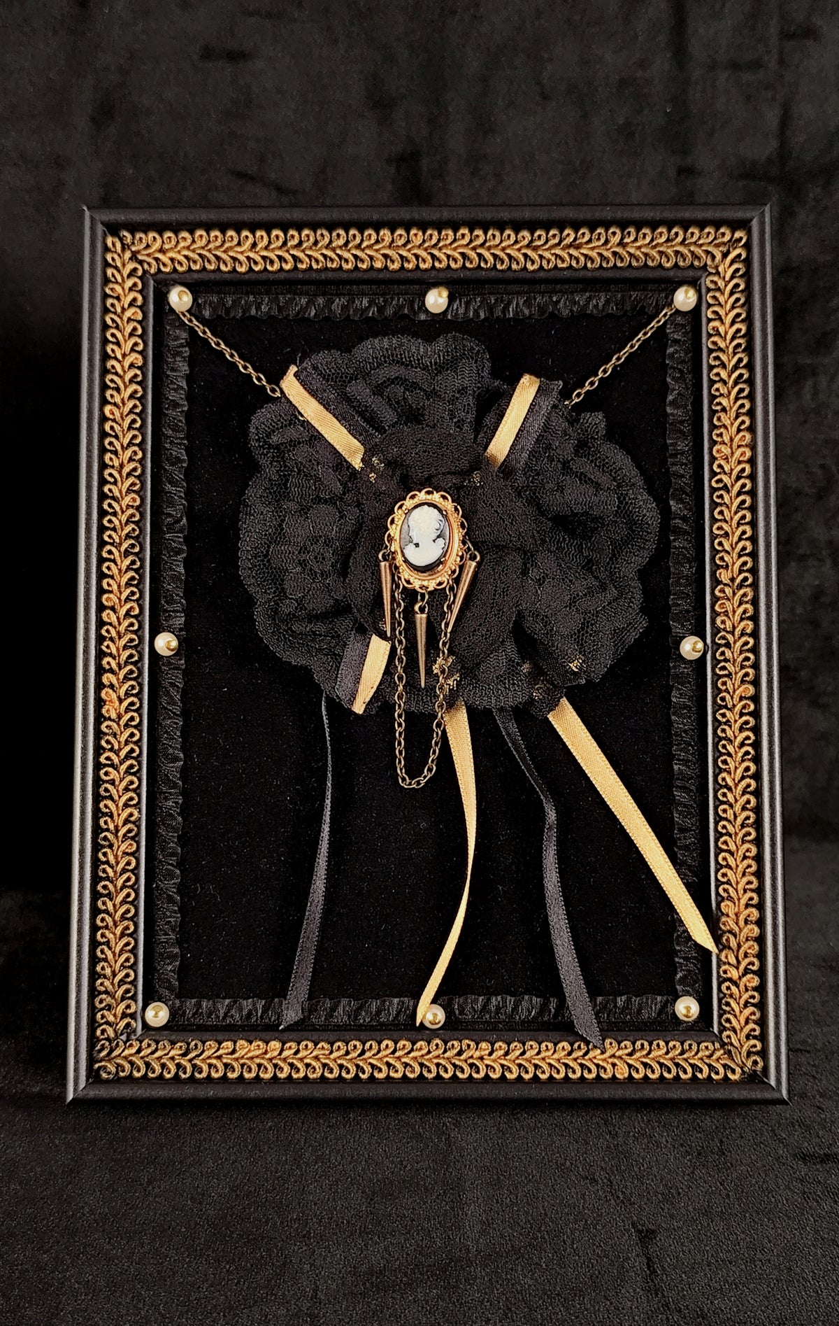 A gothic-style black lace rose brooch with a central cameo and gold chains, mounted in an ornate black and gold table frame.