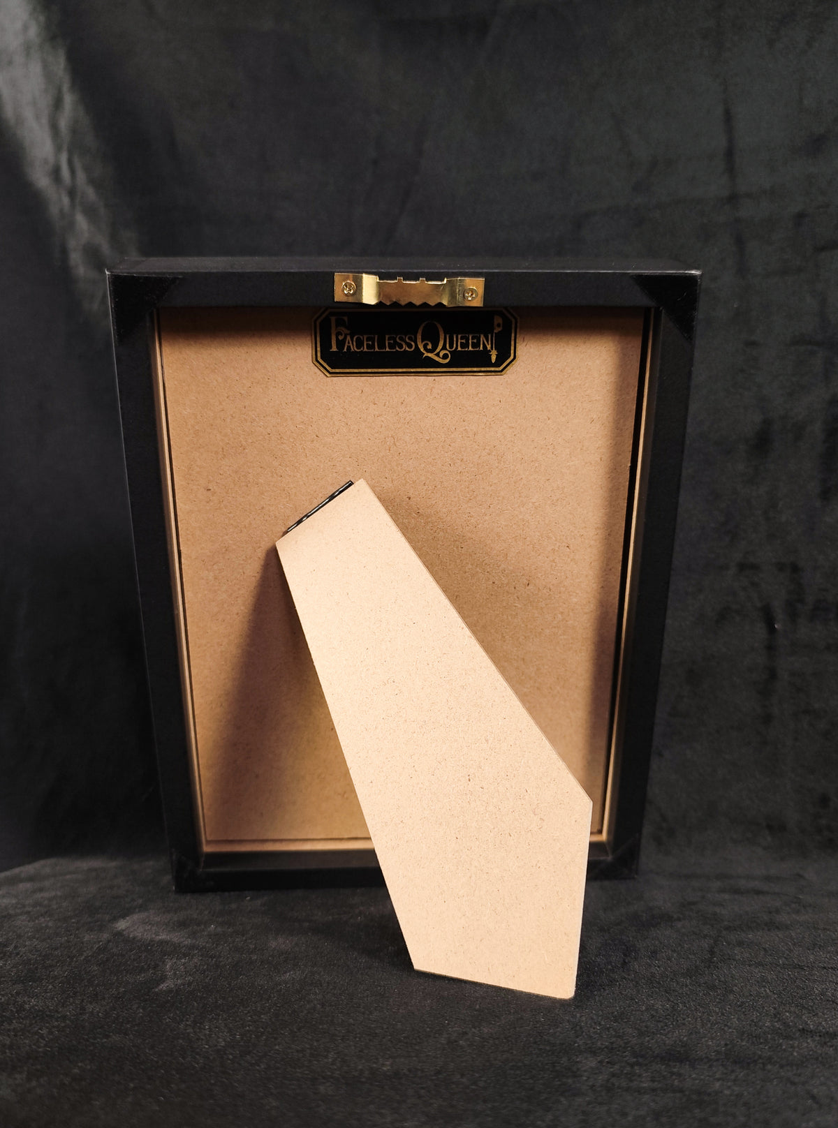 A square black picture frame with a gold-toned inner border and a blank beige backing board leaning in front, photographed against a dark textured backdrop. The brand name 'Faceless Queen' is visible on a small gold plaque at the top.