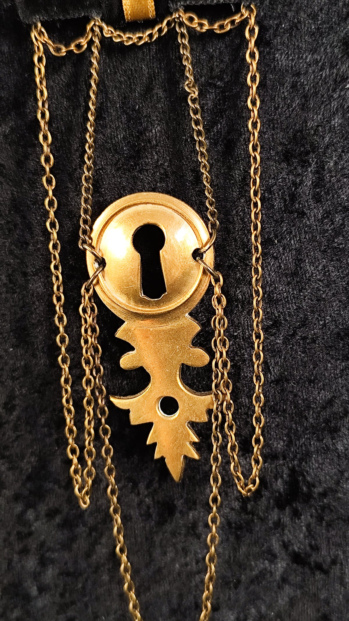 A vintage-style gold keyhole brooch and leaf-like detailing, hanging from a chain against a black textured background.