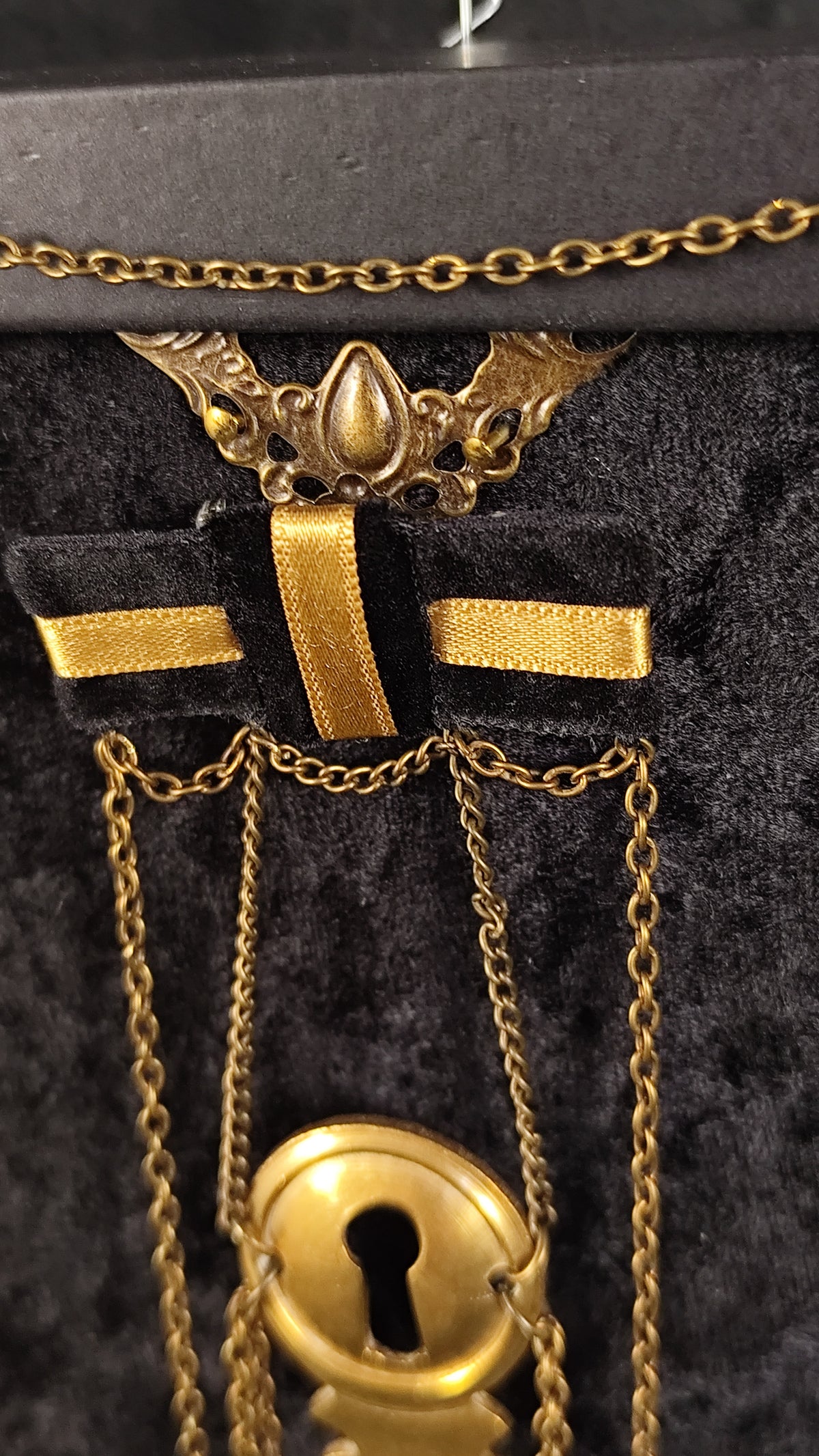 Gold-toned vintage-style chain necklace with a decorative winged top, a bow-shaped centerpiece, and a large keyhole pendant, displayed on a black velvet surface.