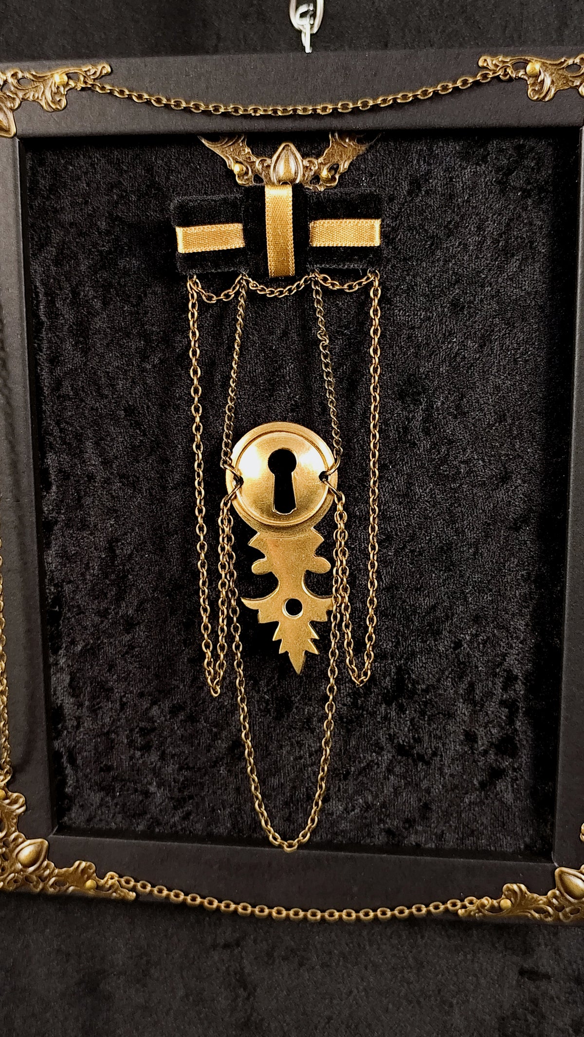 A gothic-style gold keyhole brooch displayed on a black velvet-lined presentation frame. The brooch features a gold keyhole with a leaf-shaped pendant, hanging from chains attached to a black cross with gold wings at the top.