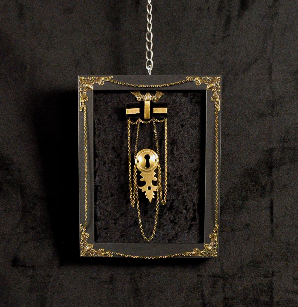 A gothic-style keyhole brooch displayed inside a black shadow box frame with ornate gold trim, hanging from a silver chain against a dark textured background.