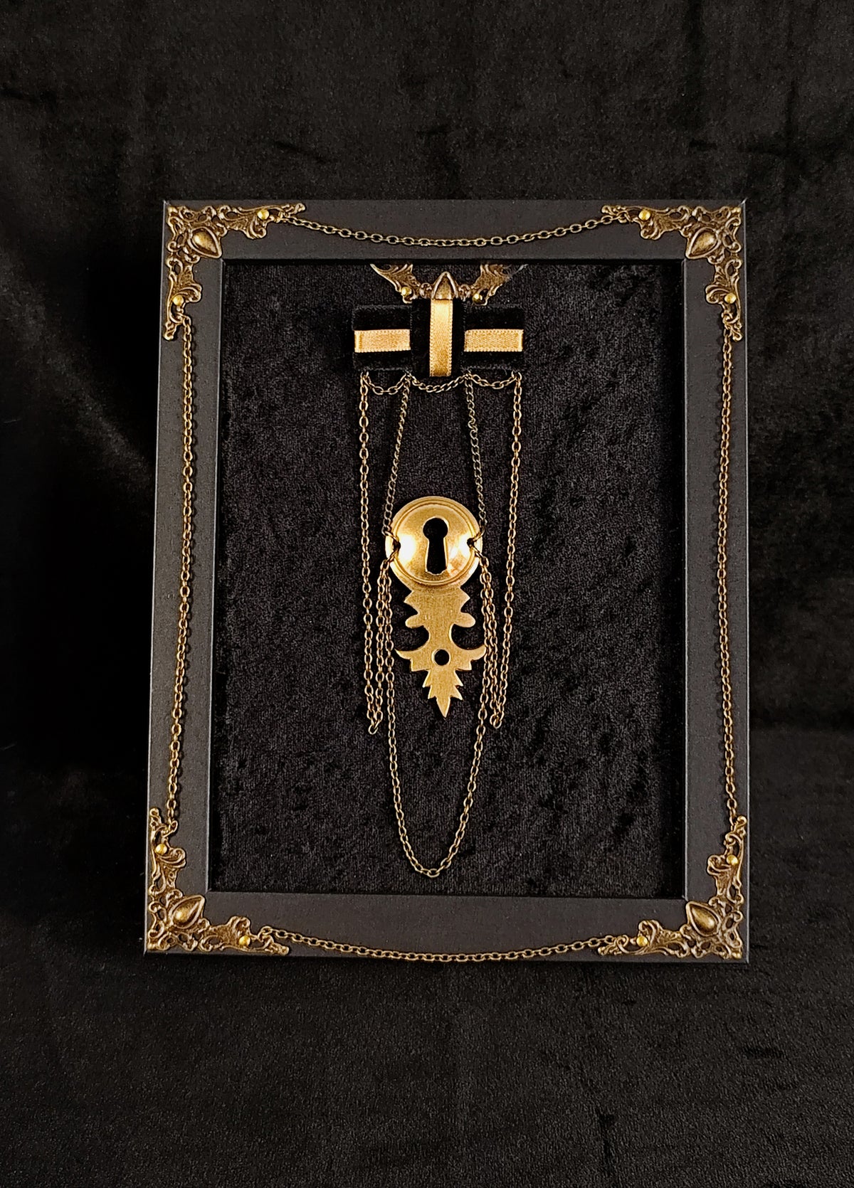 A gothic-style brooch with a keyhole and chains, displayed inside a black shadowbox frame with ornate gold corner details on a black textured background.
