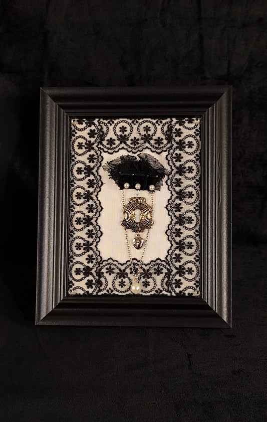A framed black and white lace art piece featuring a vintage-style keyhole and a black tulle ornament, set against a dark textured background.