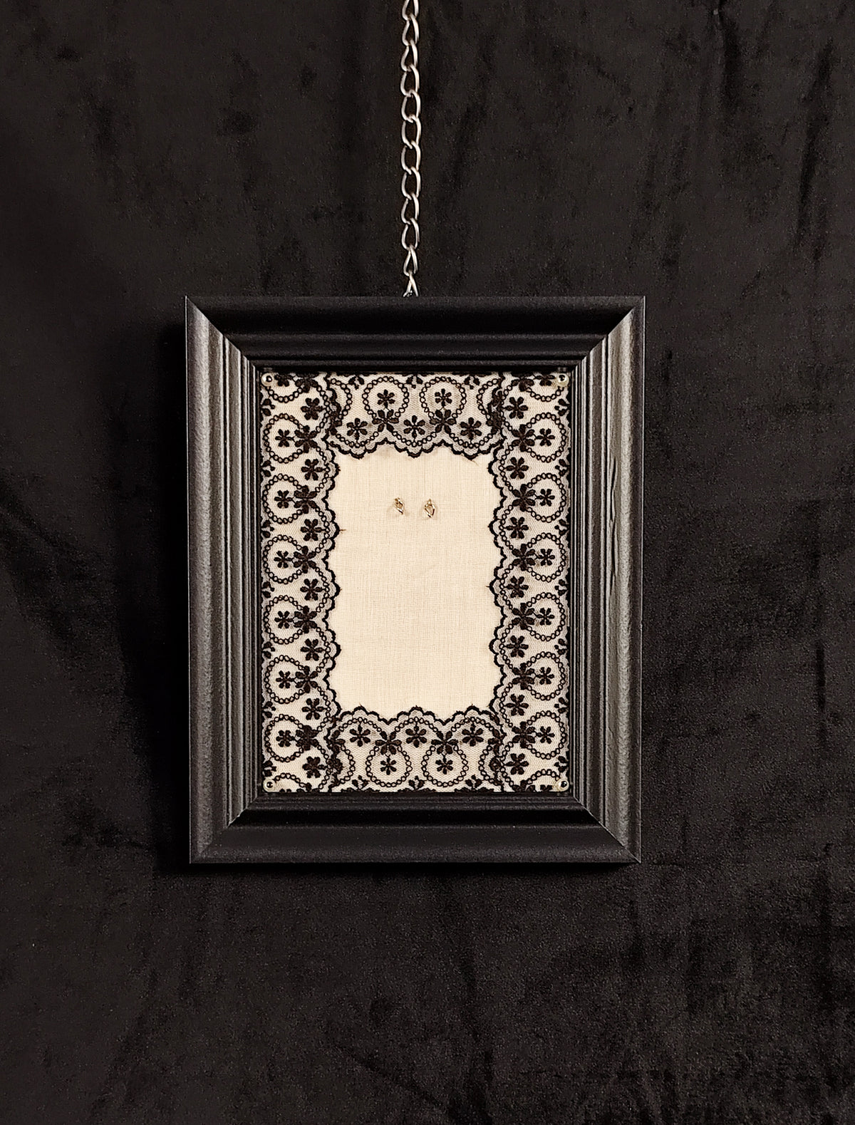 A black rectangular picture frame with an ornate black-and-cream floral lace border, hanging from a silver chain against a dark textured wall.
