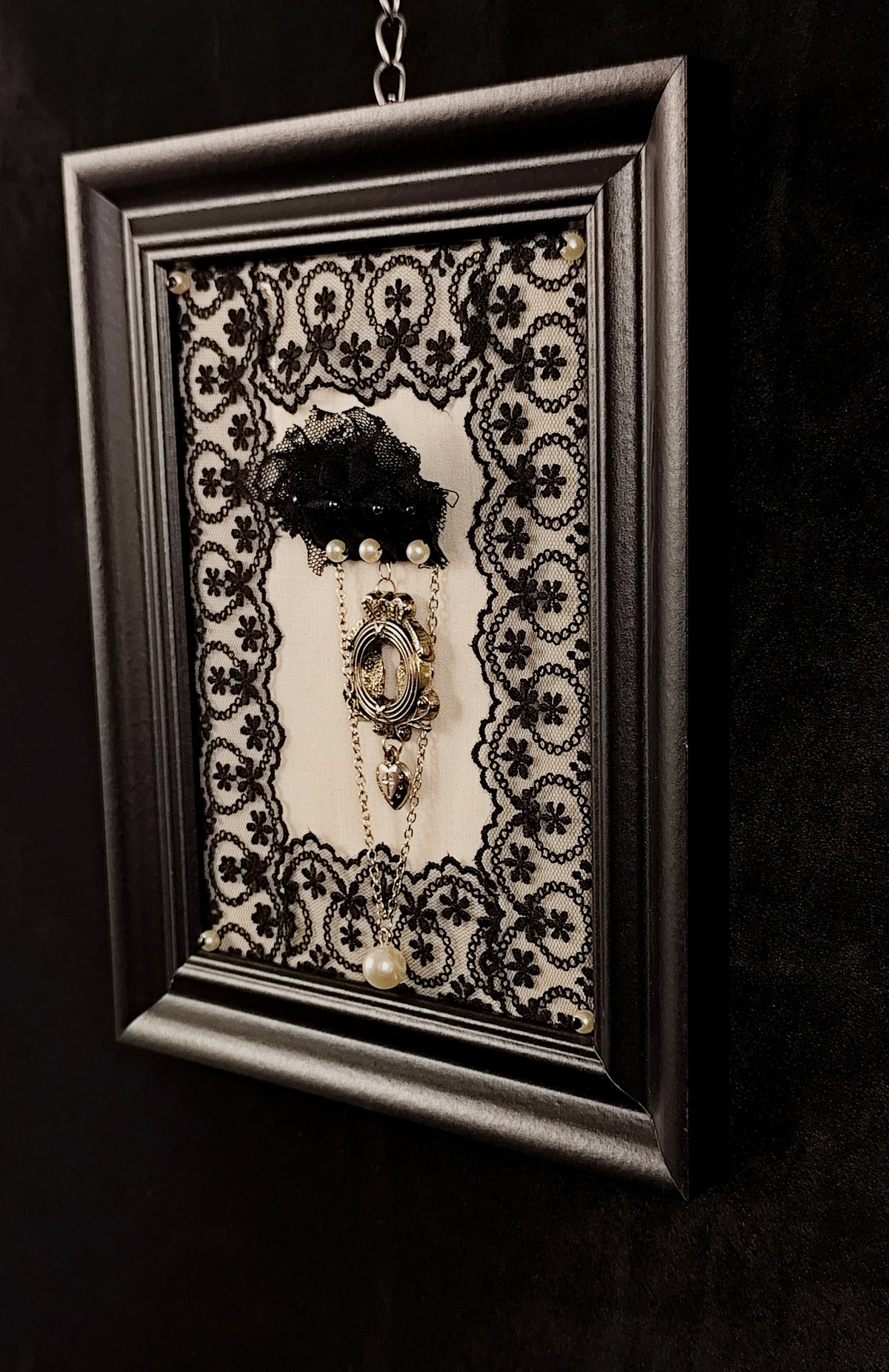 A gothic-style framed wall art piece featuring a brooch with vintage-style keyhole, agnus dei heart and a chain with a faux pearl on a black and white lace background, enclosed in a black frame hanging against a dark textured wall hanging on a dark wall. 