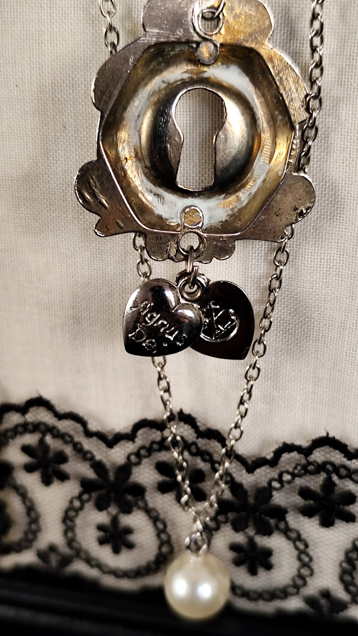 A gothic-style keyhole brooch with a silver-toned metal keyhole, an agnus dei heart, and a single white pearl at the bottom, displayed on a cream fabric background with black lace.