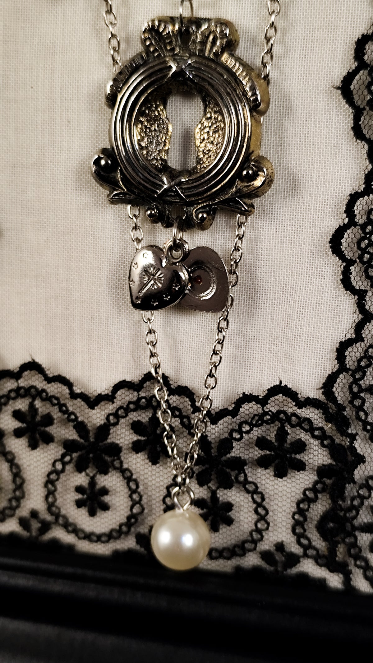 A close-up of a vintage-style brooch featuring a keyhole, an agnus dei heart, and a single pearl bead at the bottom, displayed against a textured cream fabric with black lace.