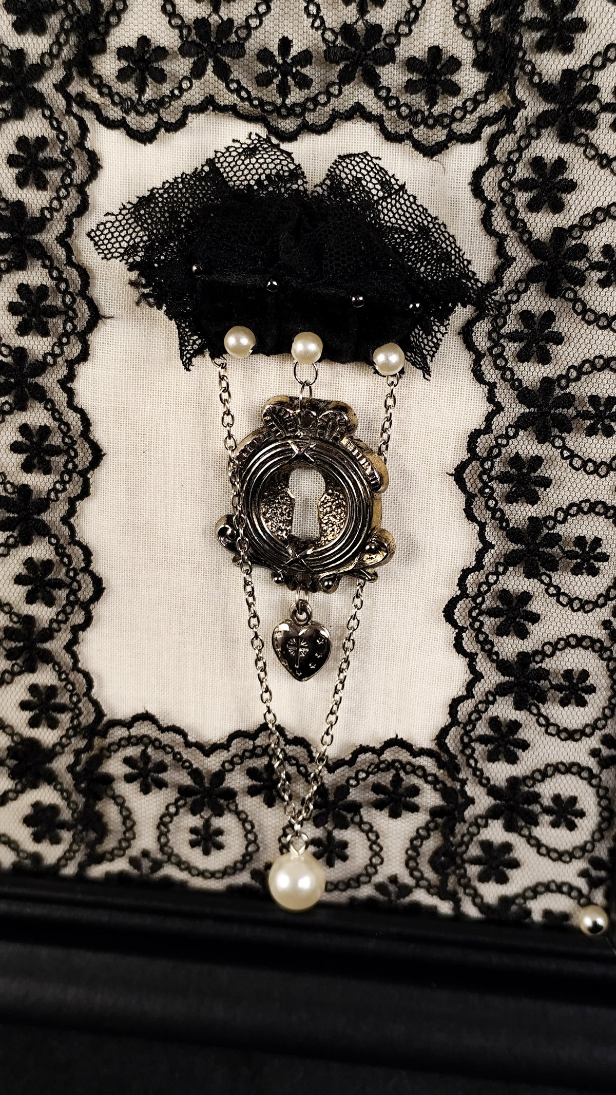 A gothic-style keyhole brooch featuring an antique silver keyhole motif, small pearl beads, and a dangling heart charm, displayed on a black lace and white fabric background.