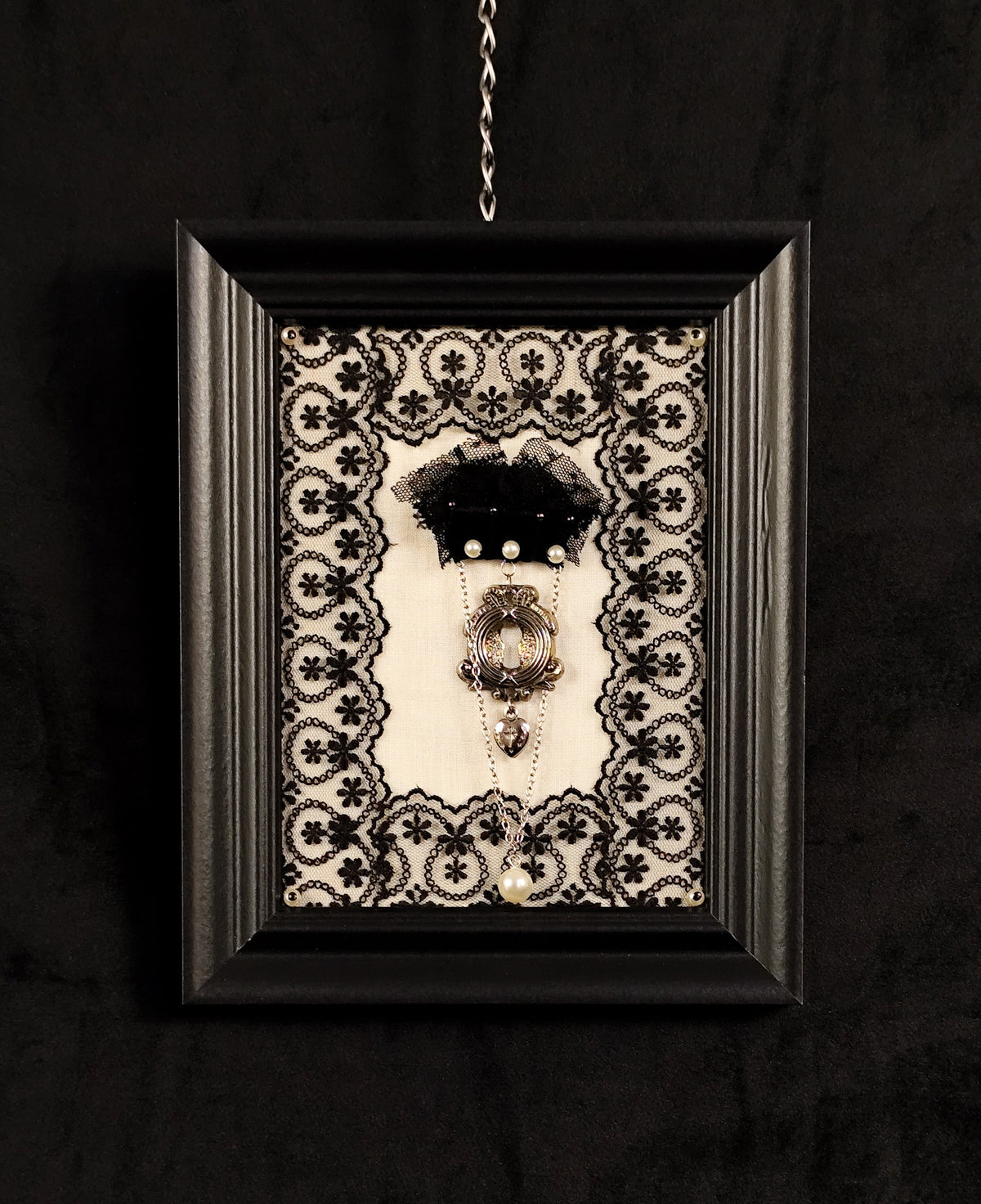 A framed gothic wall art piece featuring a brooch with vintage-style keyhole, agnus dei heart and a chain with a faux pearl on a black and white lace background, enclosed in a black frame hanging against a dark textured wall.
