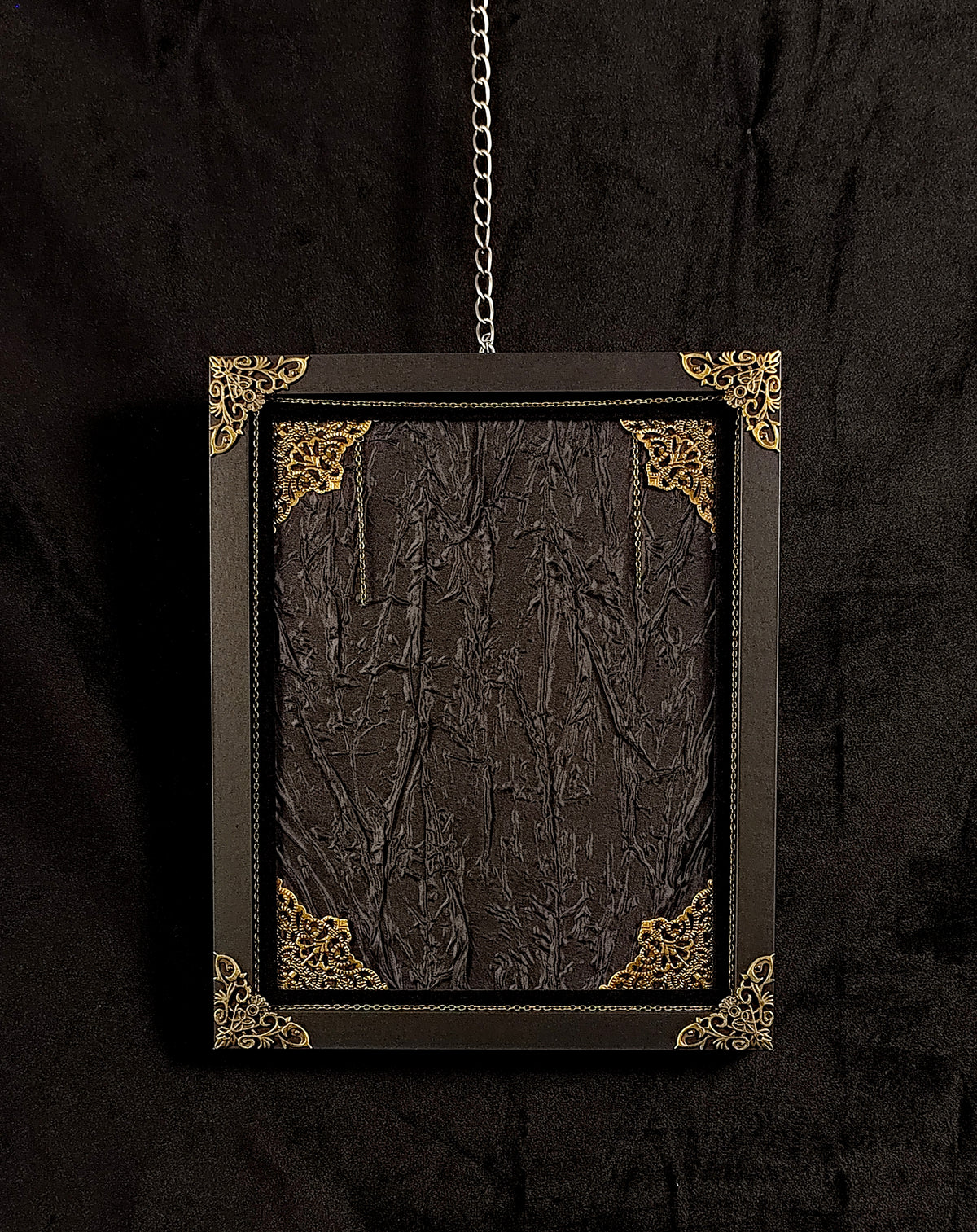 A black hanging picture frame with ornate gold corner accents and a textured black fabric, suspended by a metal chain against a black fabric backdrop.