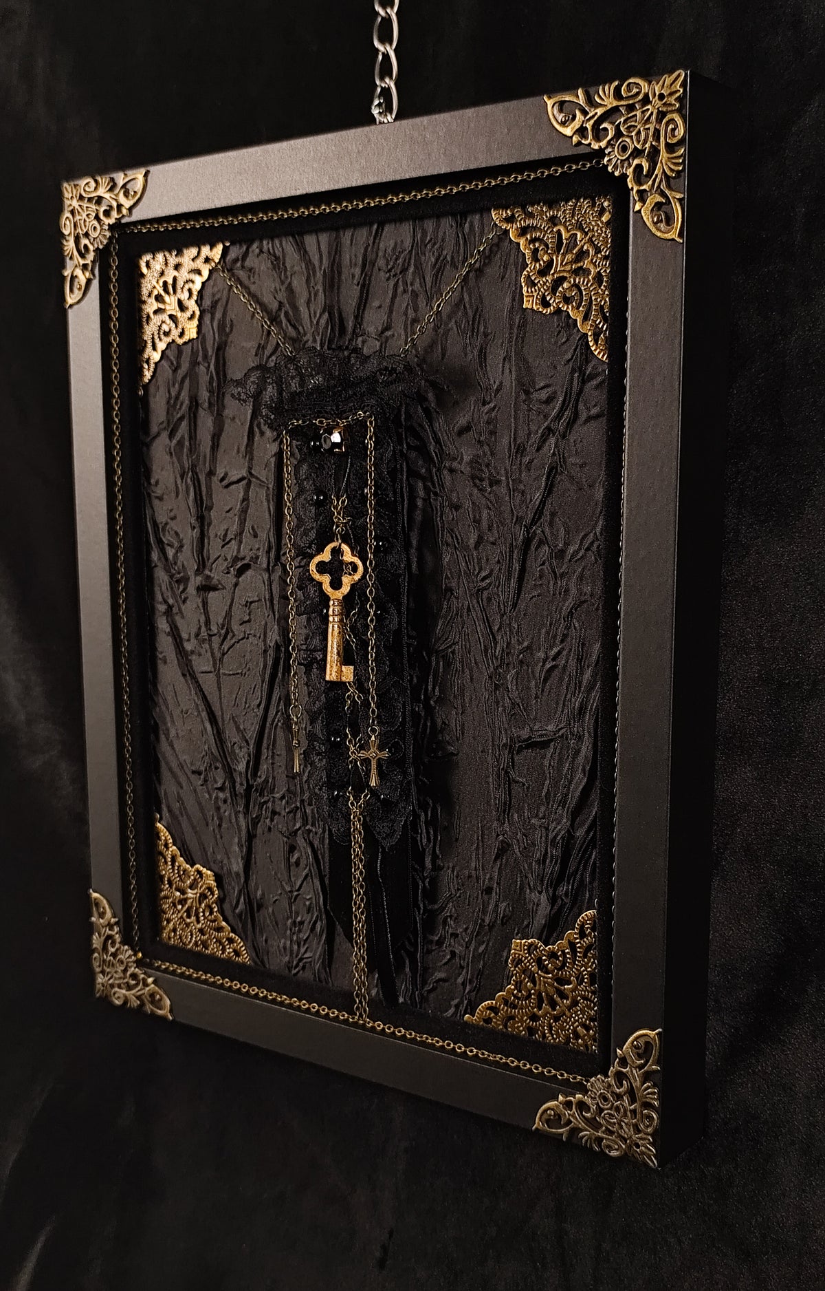 A gothic-style framed wall hanging featuring a textured black fabric with ornate gold corner details, a hanging brooch made of velvet, lace, featuring a key, chains and crosses, suspended against a dark background. Gothic home decor.