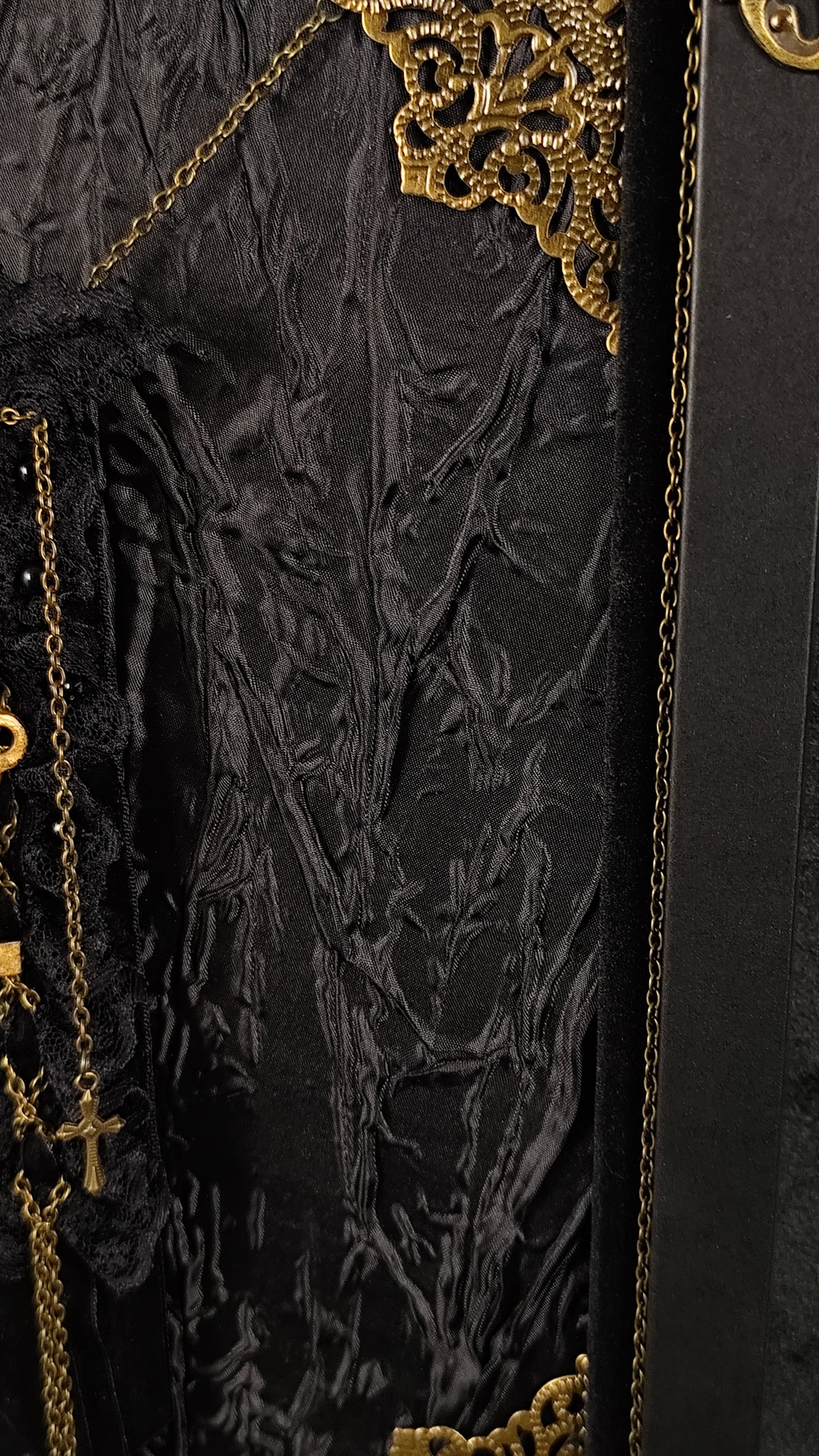 Close-up of a black textured fabric on a frame with ornate gold embroidery, chain details, and a small gold cross pendant.