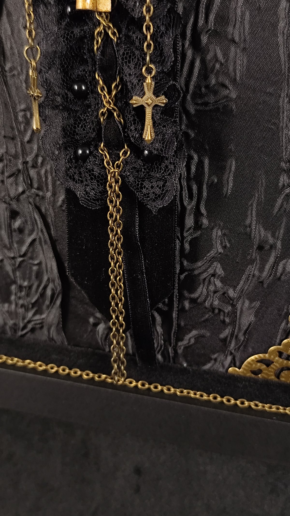 Close-up of a brooch featuring two golden crosses and chains, displayed against a black lace and textured fabric backdrop.