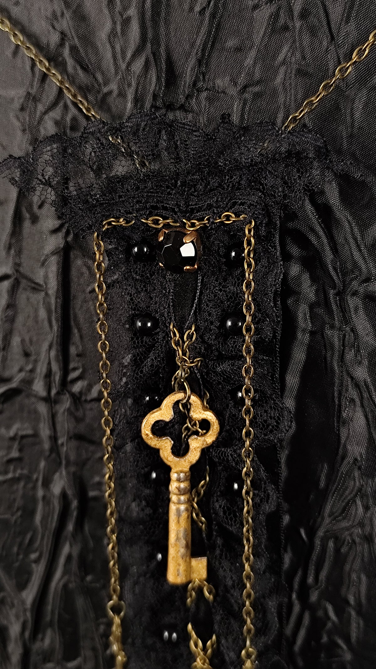 A close-up of a black lace and tulle brooch with draped gold chains and a vintage-style gold key hanging at the center, set against a dark textured fabric background.