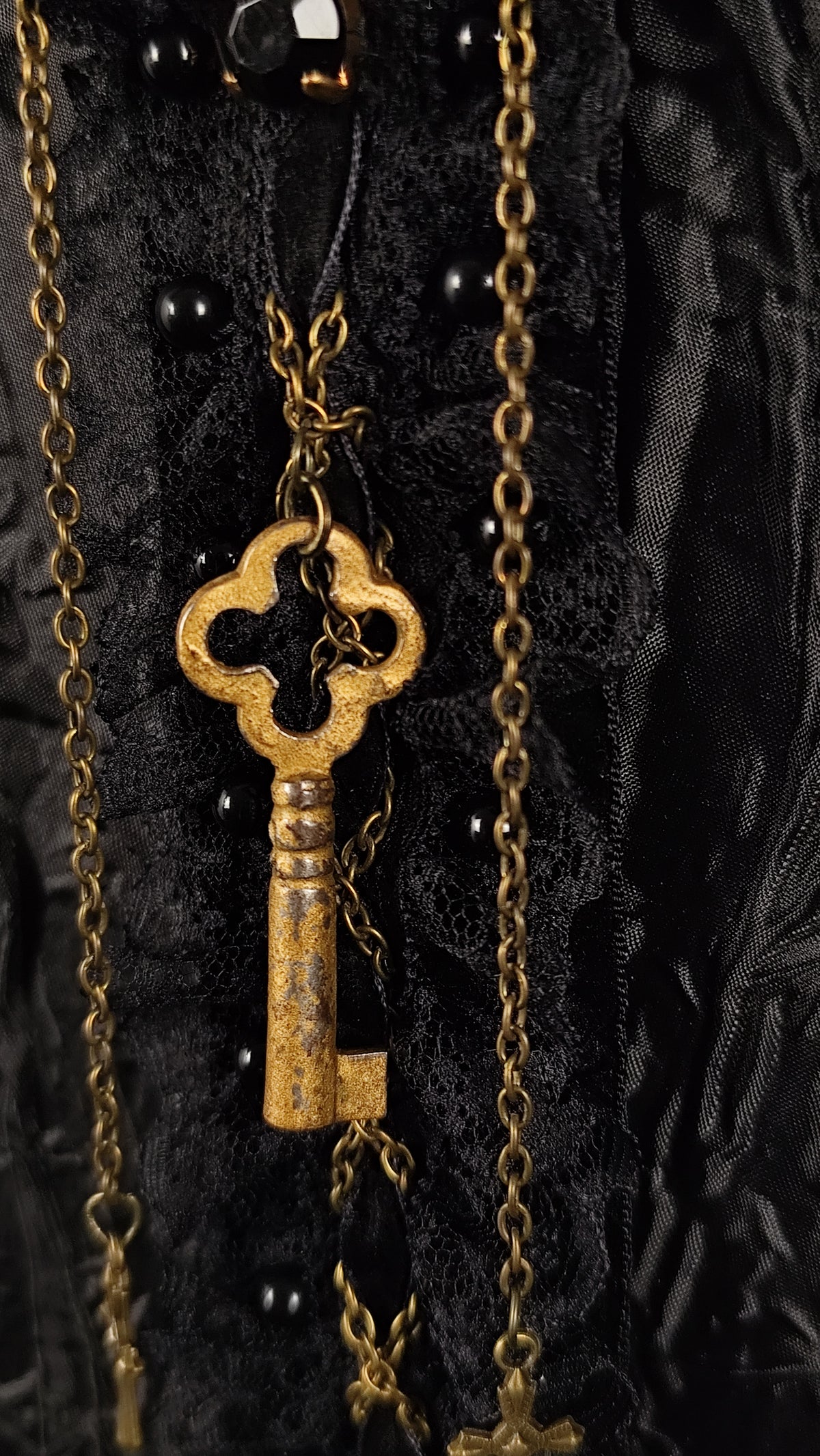 An ornate antique-style gold key hanging from a black lace and velvet garment with black beads and layered gold chains.