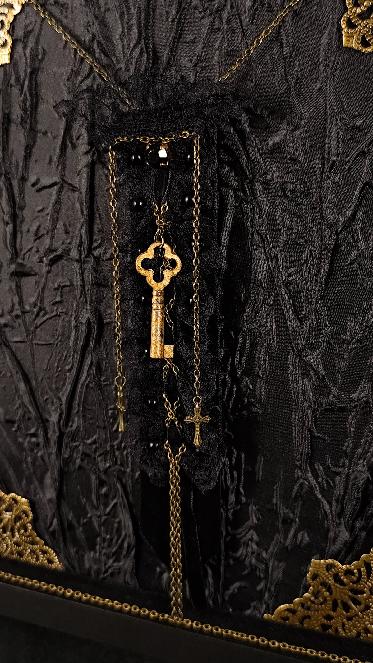 A gothic-style black lace and tulle brooch with gold chain detailing, a large ornate gold key pendant, and a small gold cross, displayed against a textured black fabric backdrop.