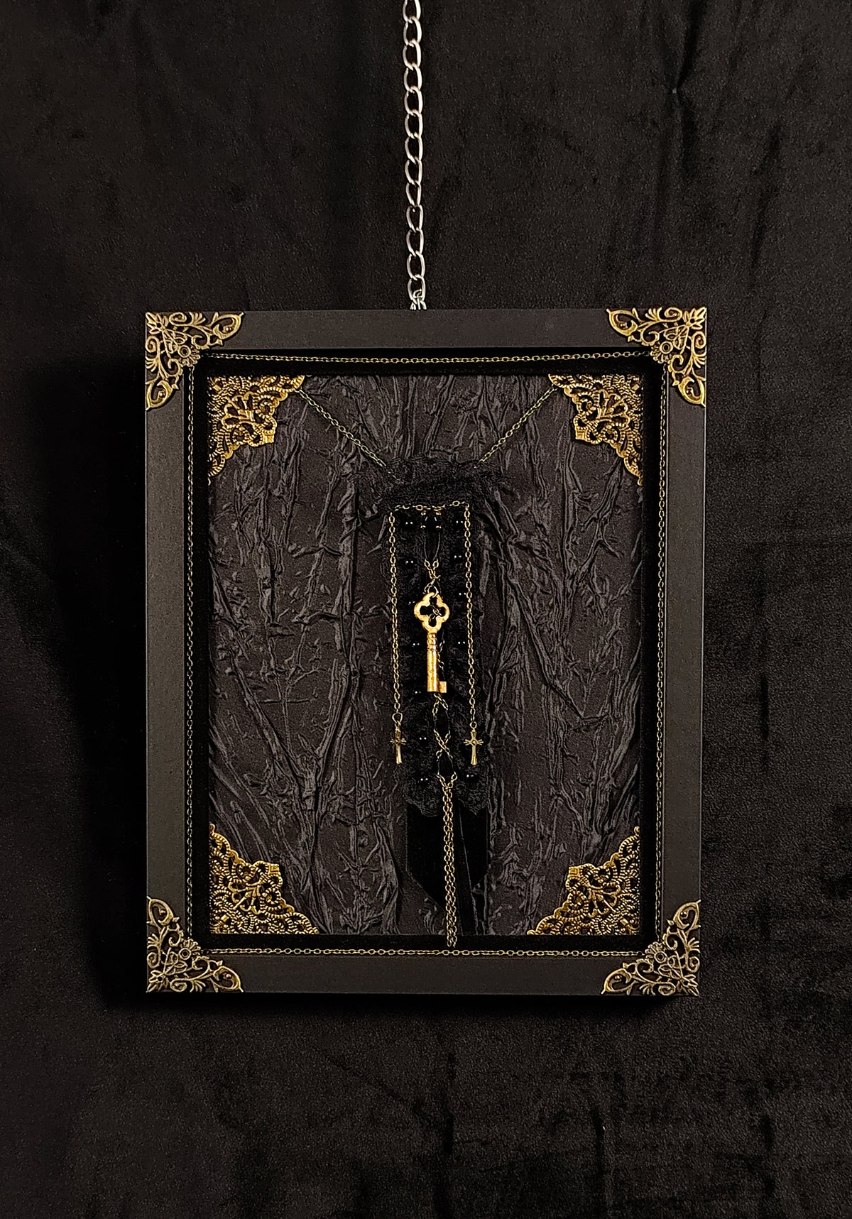 A gothic-style framed wall hanging featuring a textured black fabric with ornate gold corner details, a hanging brooch made of velvet, lace, featuring a key, chains and crosses, suspended against a dark background.