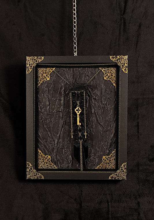 A gothic-style framed wall hanging featuring a textured black fabric with ornate gold corner details, a hanging brooch made of velvet, lace, featuring a key, chains and crosses, suspended against a dark background.