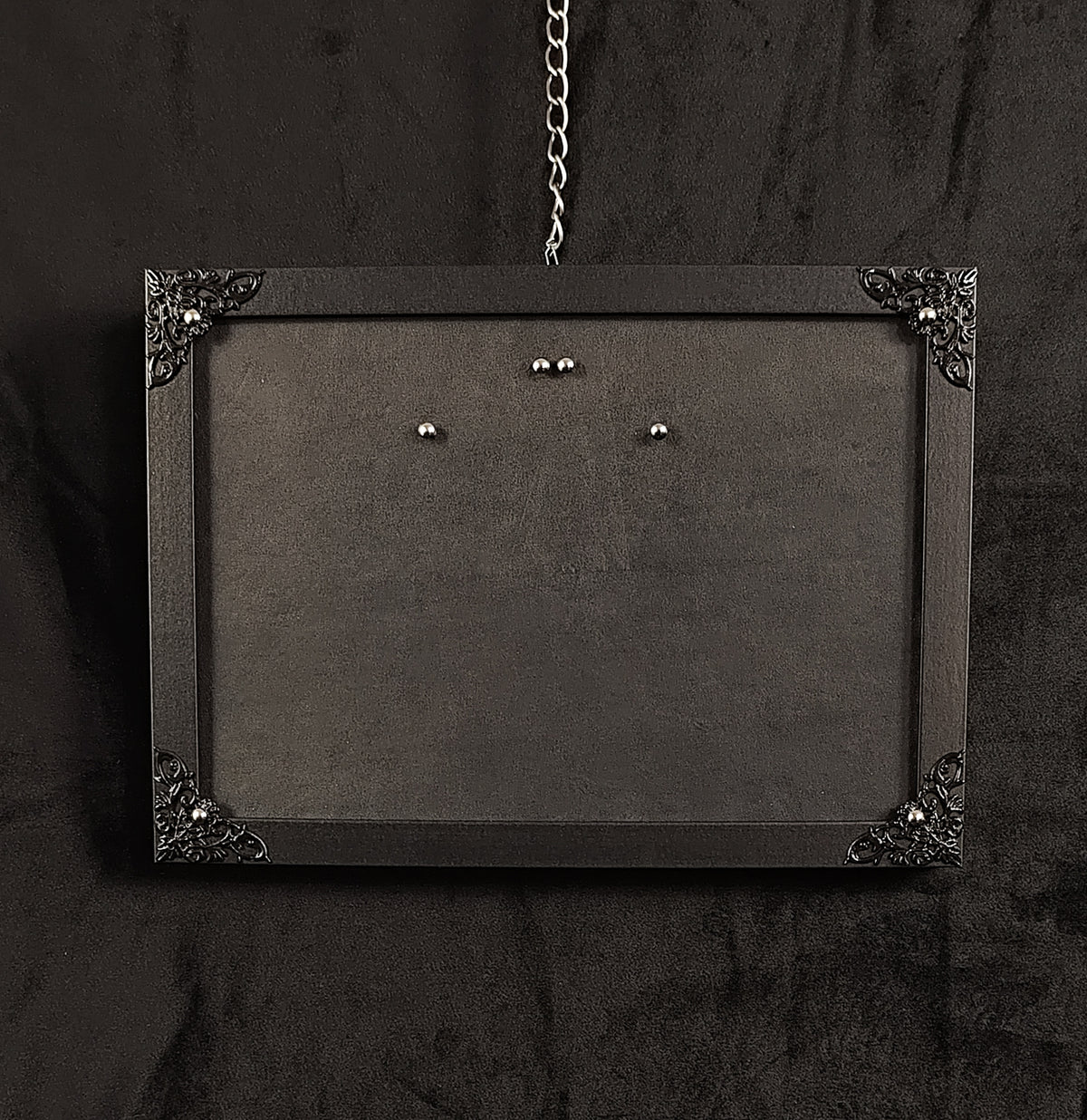 A black rectangular wall-mounted picture frame with ornate metal corner details and a hanging chain, shown against a dark textured background.