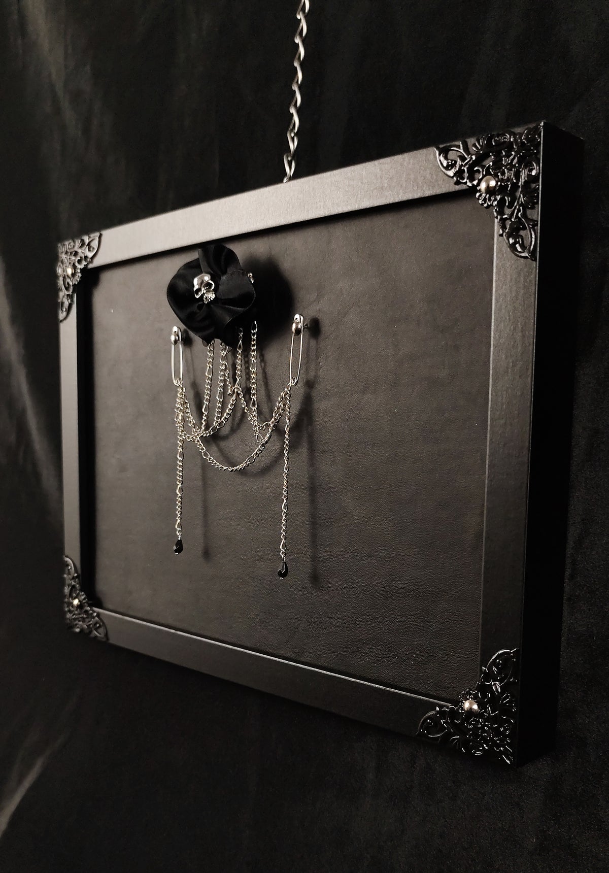 A black gothic-style framed wall display with ornate metal corner details, holding a black flower-shaped brooch or pin with dangling silver chains against a dark background.