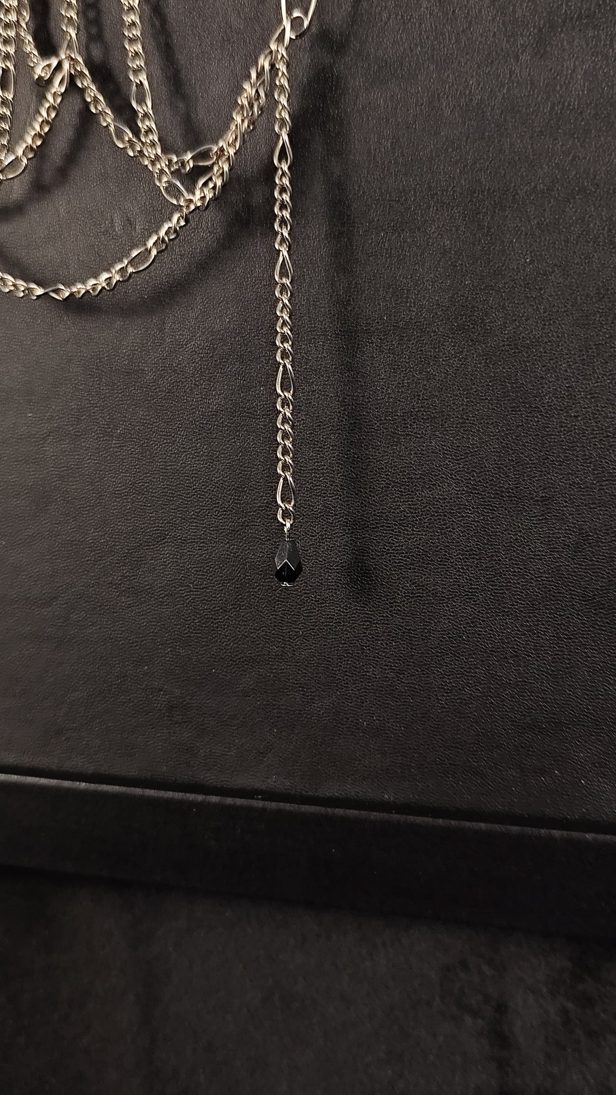 A close-up of a silver-toned chain with a long drop chain ending in a small black bead, displayed against a dark textured background.