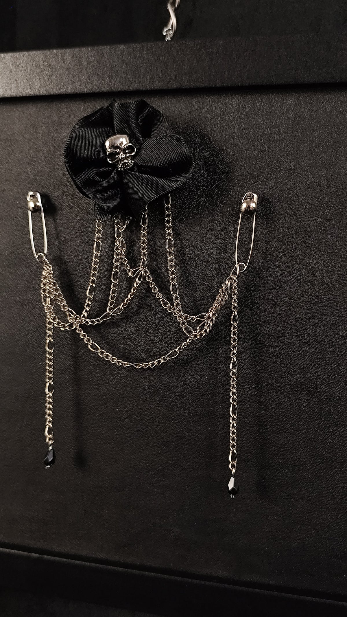 A gothic-style metal brooch or pin featuring two silver safety pins connected by draped chains, with a black satin flower adorned with a small silver skull, displayed on a black background.