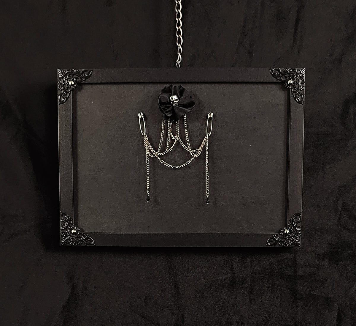 A black gothic-style hanging display board with ornate corner details, featuring a brooch made with black fabric flower with a skull center and silver chain accessories hanging from safety pins.