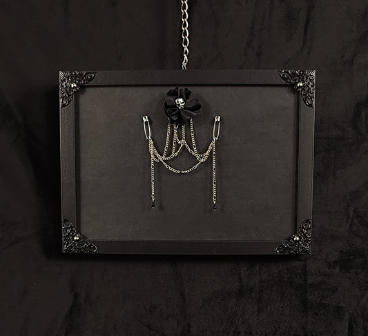 A black gothic-style hanging display board with ornate corner details, featuring a brooch made with black fabric flower with a skull center and silver chain accessories hanging from safety pins.