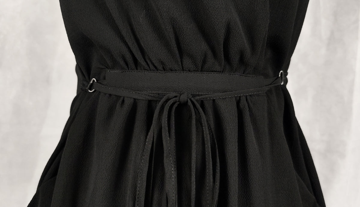Close-up of a black dress with a matching fabric tie belt and small metal ring details at the waist, shown against a light gray background.
