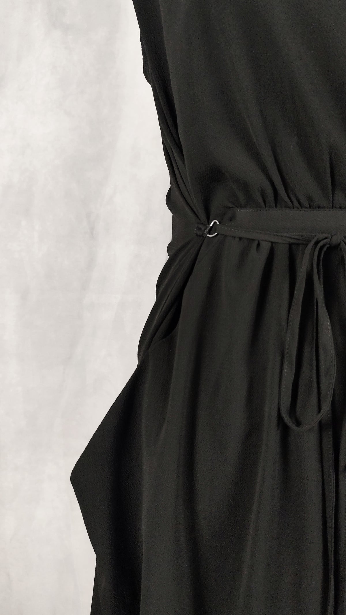 Close-up of a black wrap-style dress with a tie belt and small metal ring detail at the waist, shown against a light gray textured background.