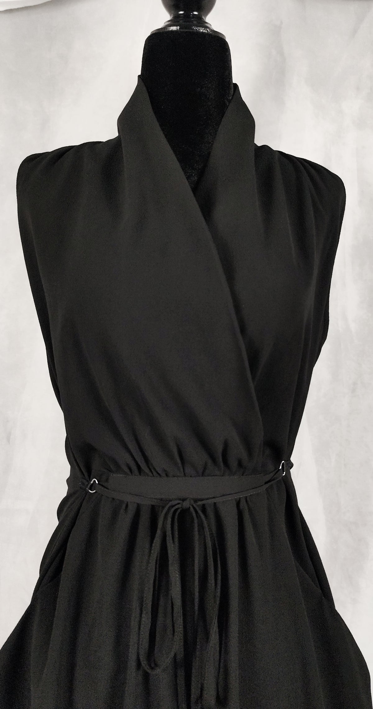 A black sleeveless wrap-style dress displayed on a black mannequin torso against a light gray textured background.