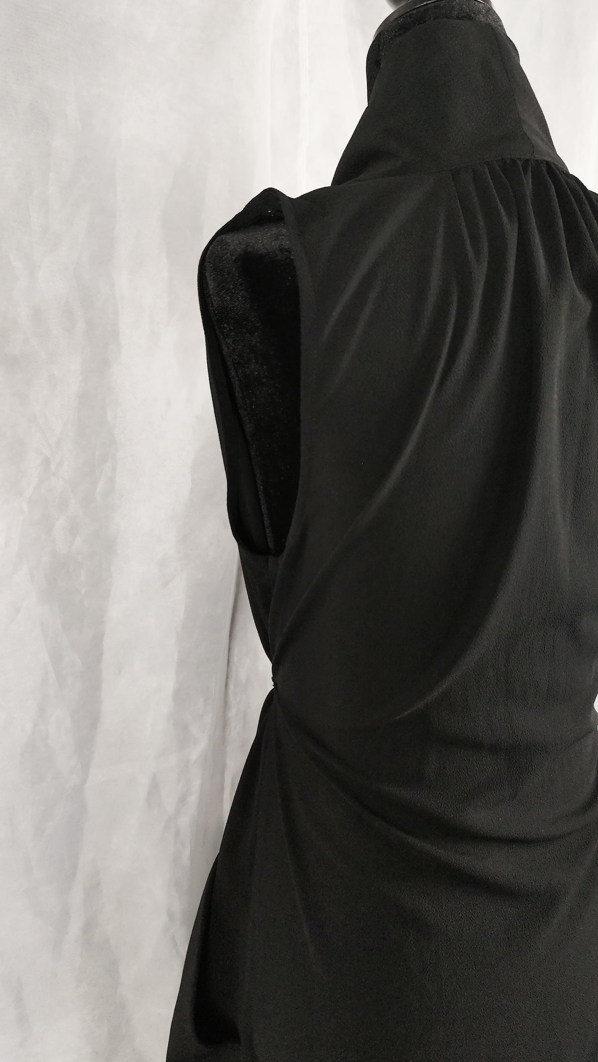 A close-up of a black sleeveless top or dress with a gathered, draped back design, displayed on a mannequin against a light gray textured backdrop.