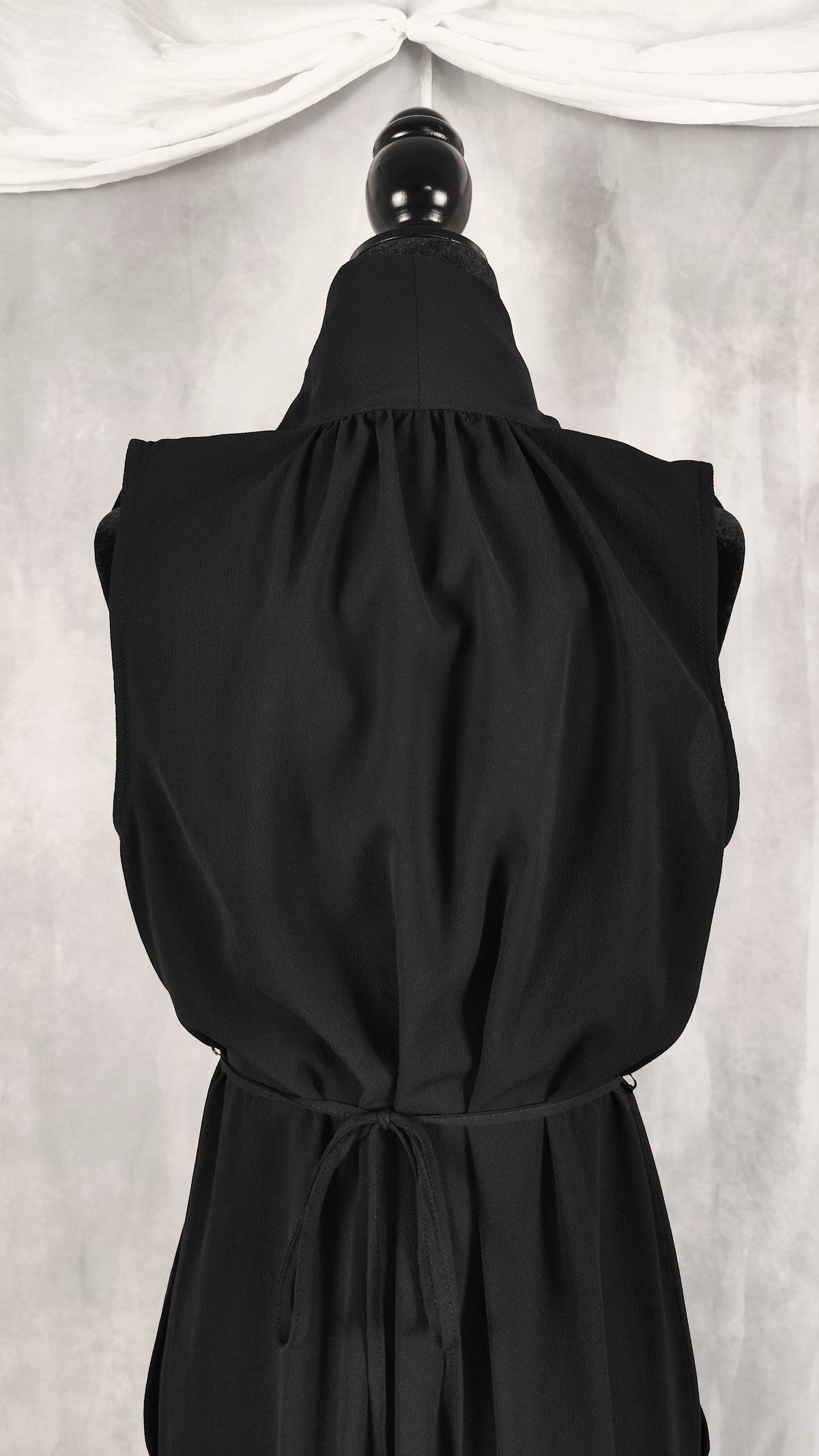 A black sleeveless dress with a high gathered neckline and a drawstring waist, displayed on a black dress form against a light gray backdrop with draped white fabric at the top.