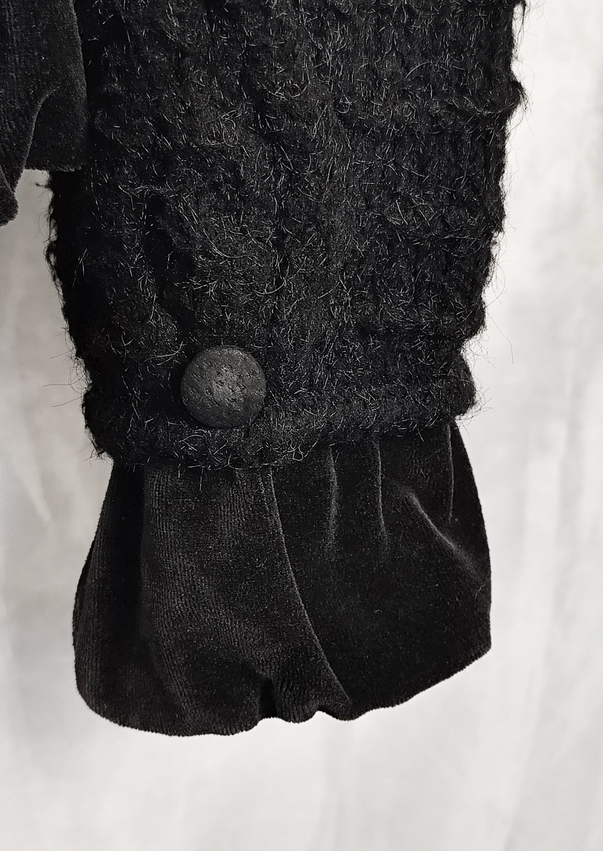 Close-up of a black textured knit sleeve with a single round button and a gathered black velvet cuff, shown against a light neutral background.