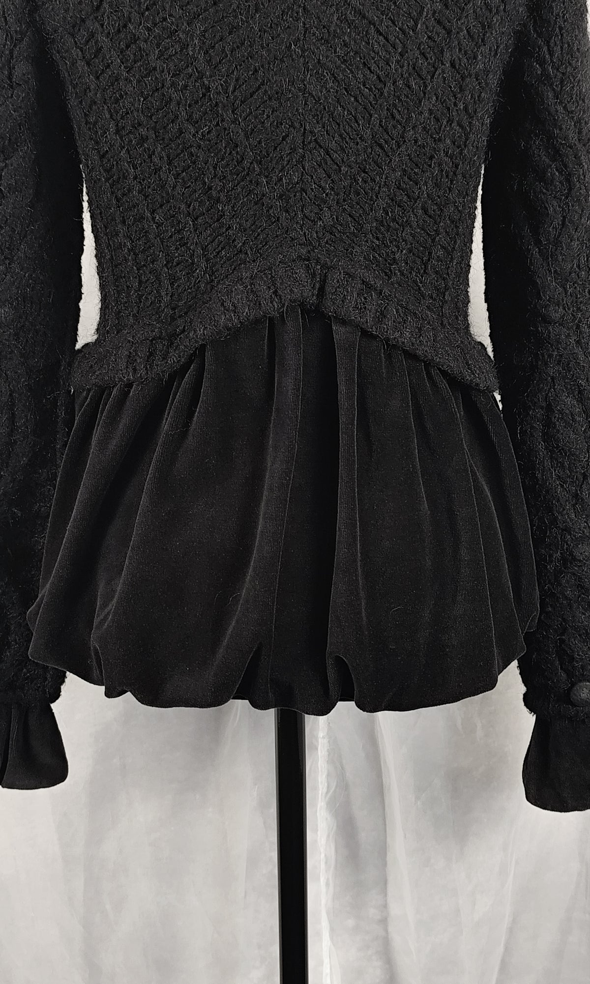 A close-up of a black textured knit jacket with long sleeves and a gathered, ruffled velvet-like hem, displayed on a mannequin against a light gray background.