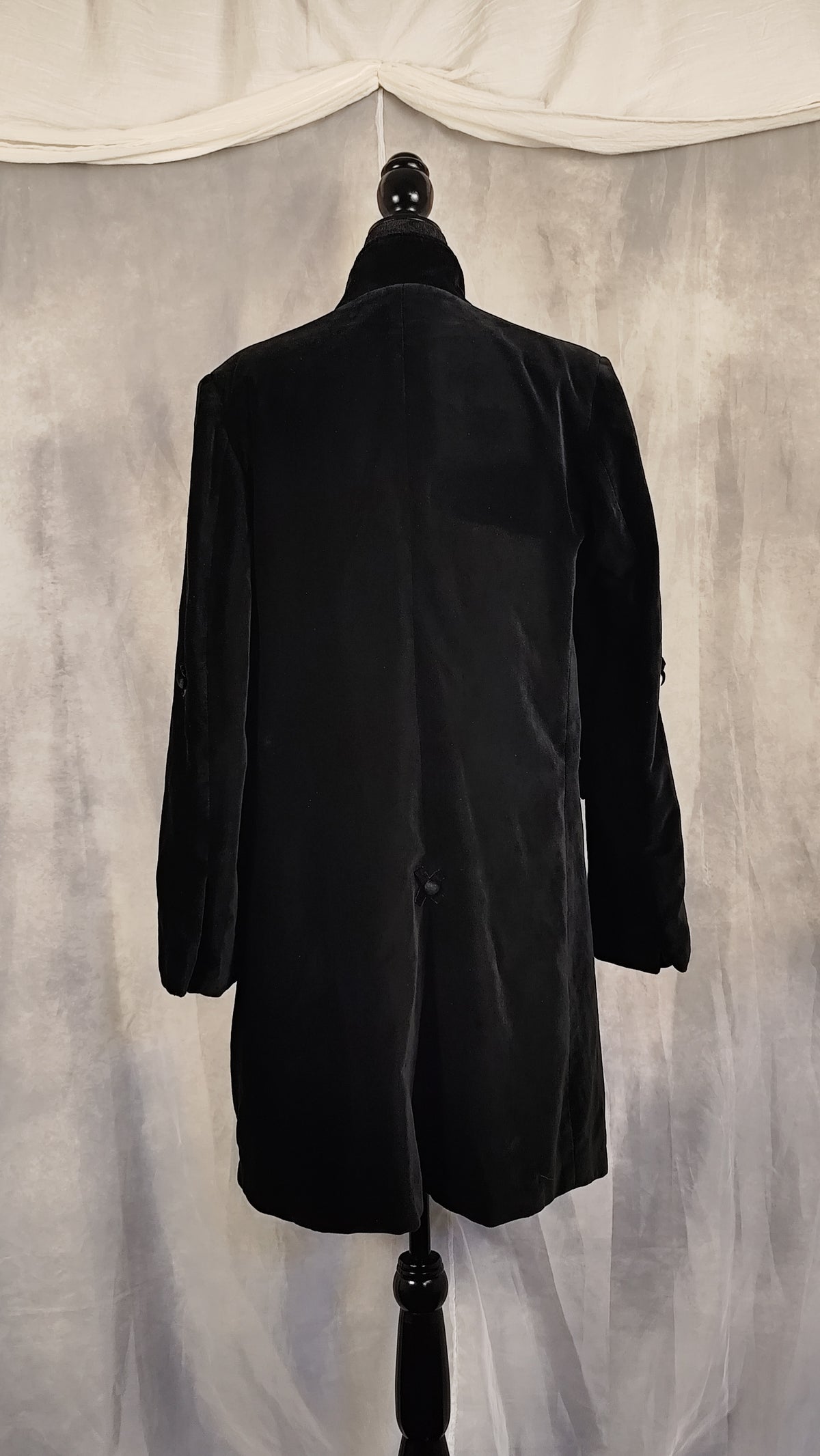 A black velvet long coat displayed on a black dress form mannequin against a gray draped fabric backdrop.