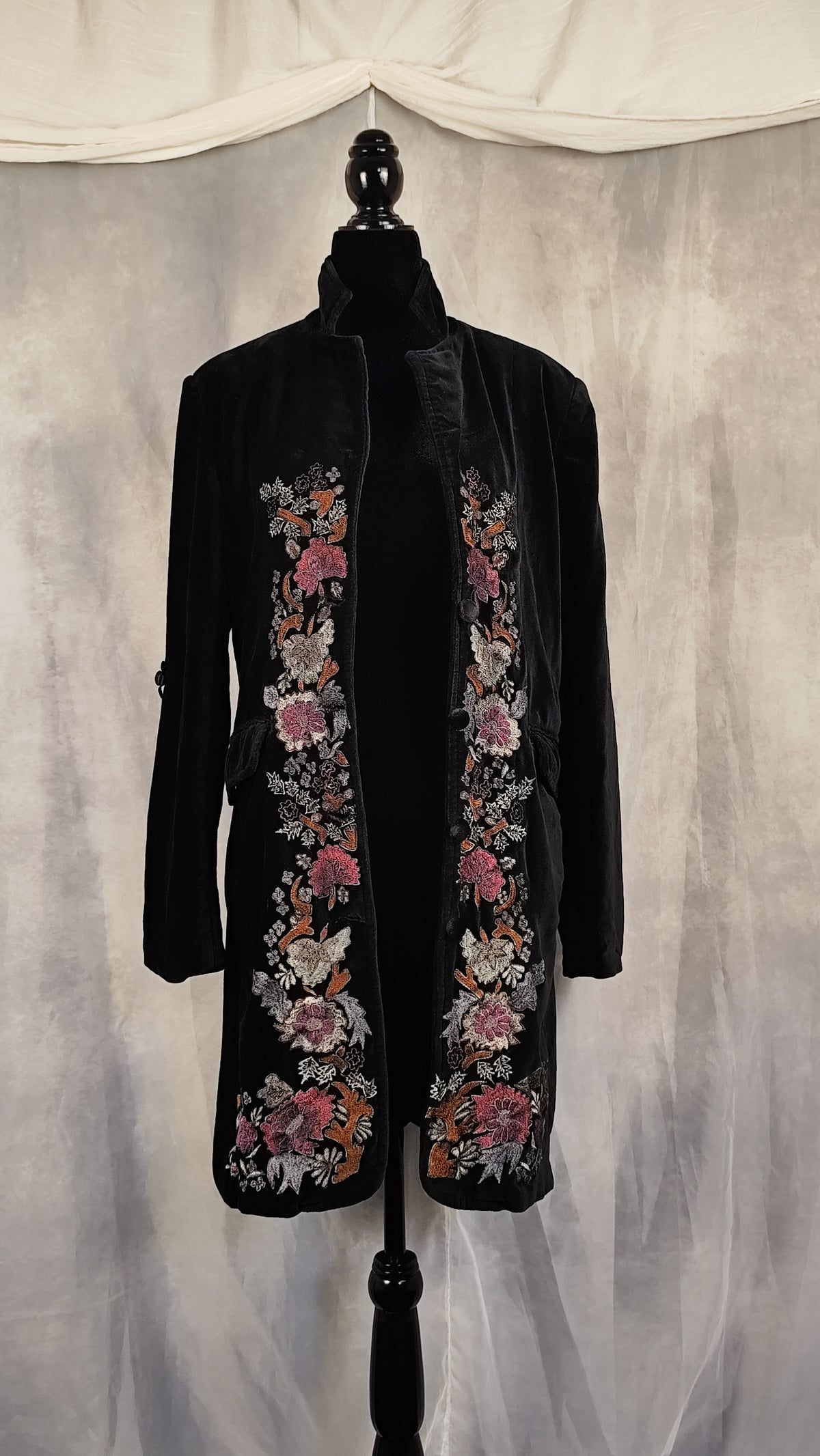 A black velvet longline coat with intricate floral embroidery in pink, white, and orange, displayed on a black dress form mannequin against a draped gray fabric backdrop.