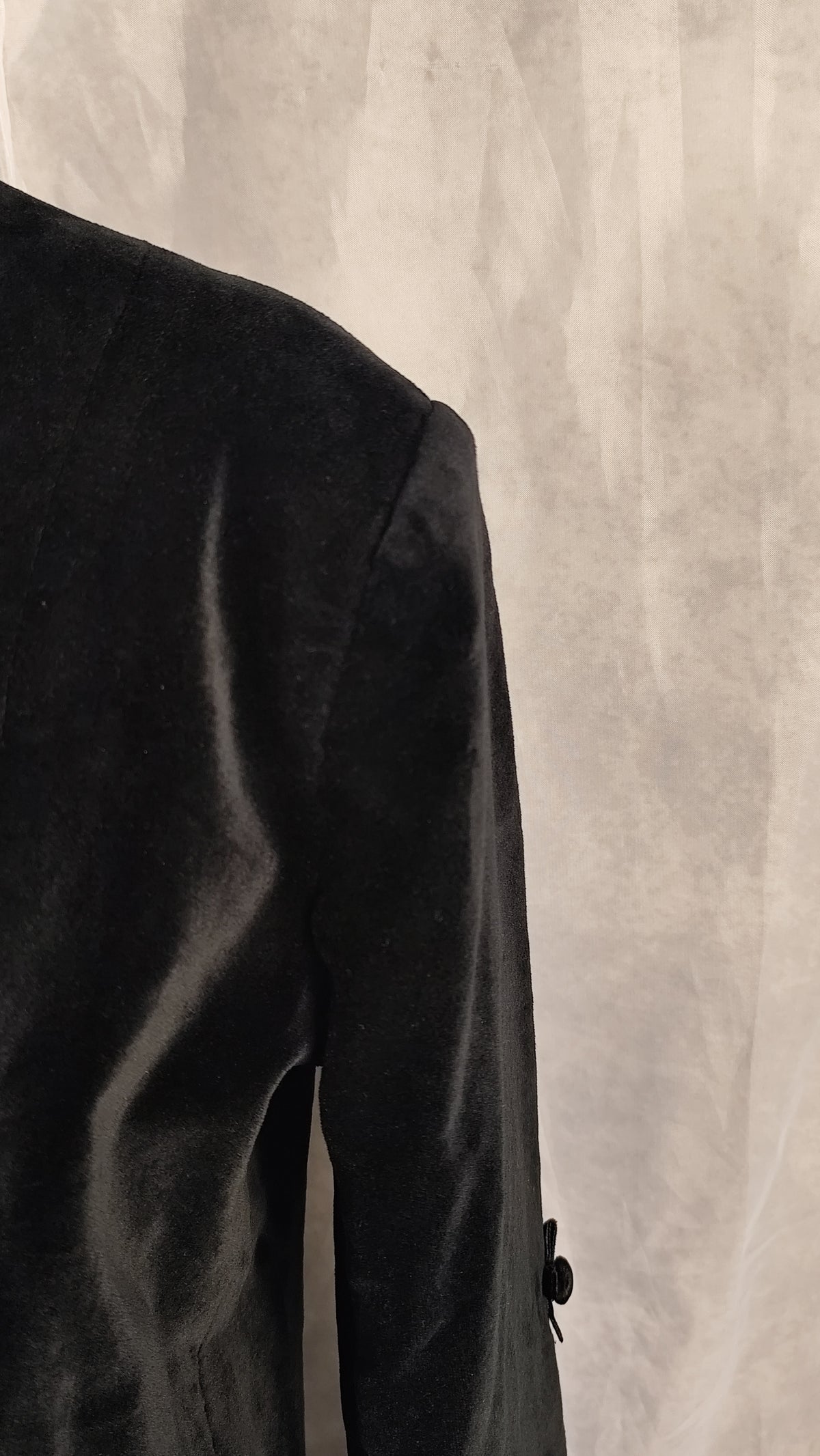 Close-up of a black velvet coat sleeve and shoulder, shown against a textured light gray background.