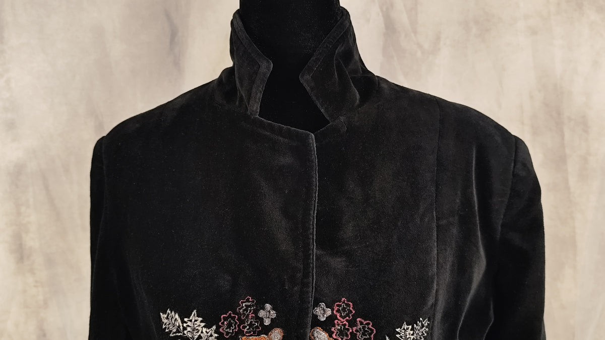 A black velvet coat with a high stand collar and embroidered floral details at the bottom, displayed on a black mannequin against a textured beige background.