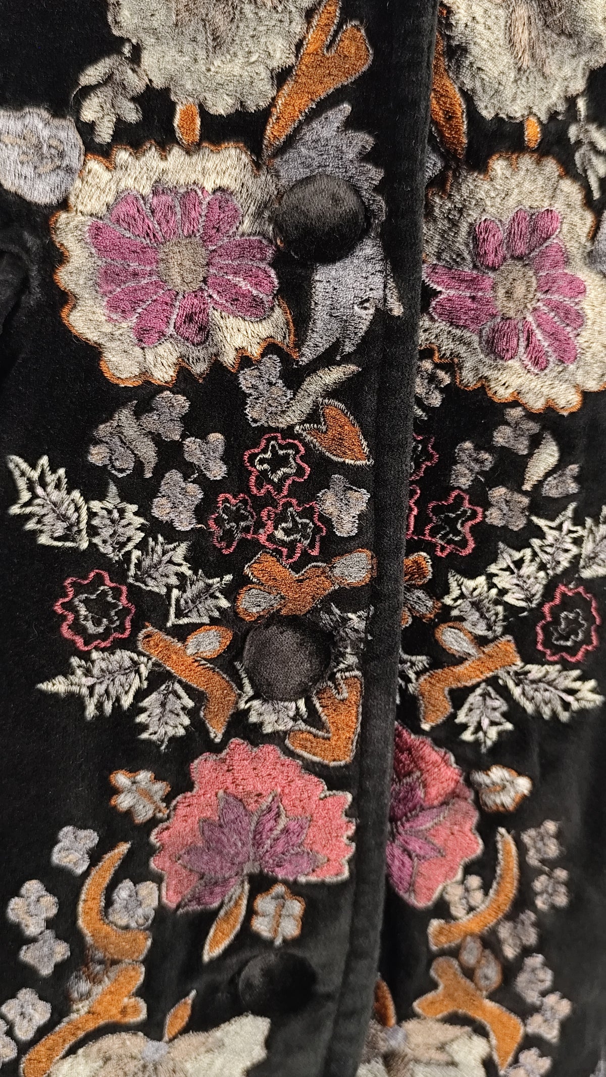 Close-up of a black embroidered jacket or vest featuring intricate floral patterns in pink, orange, gray, and white, with dark buttons down the front.