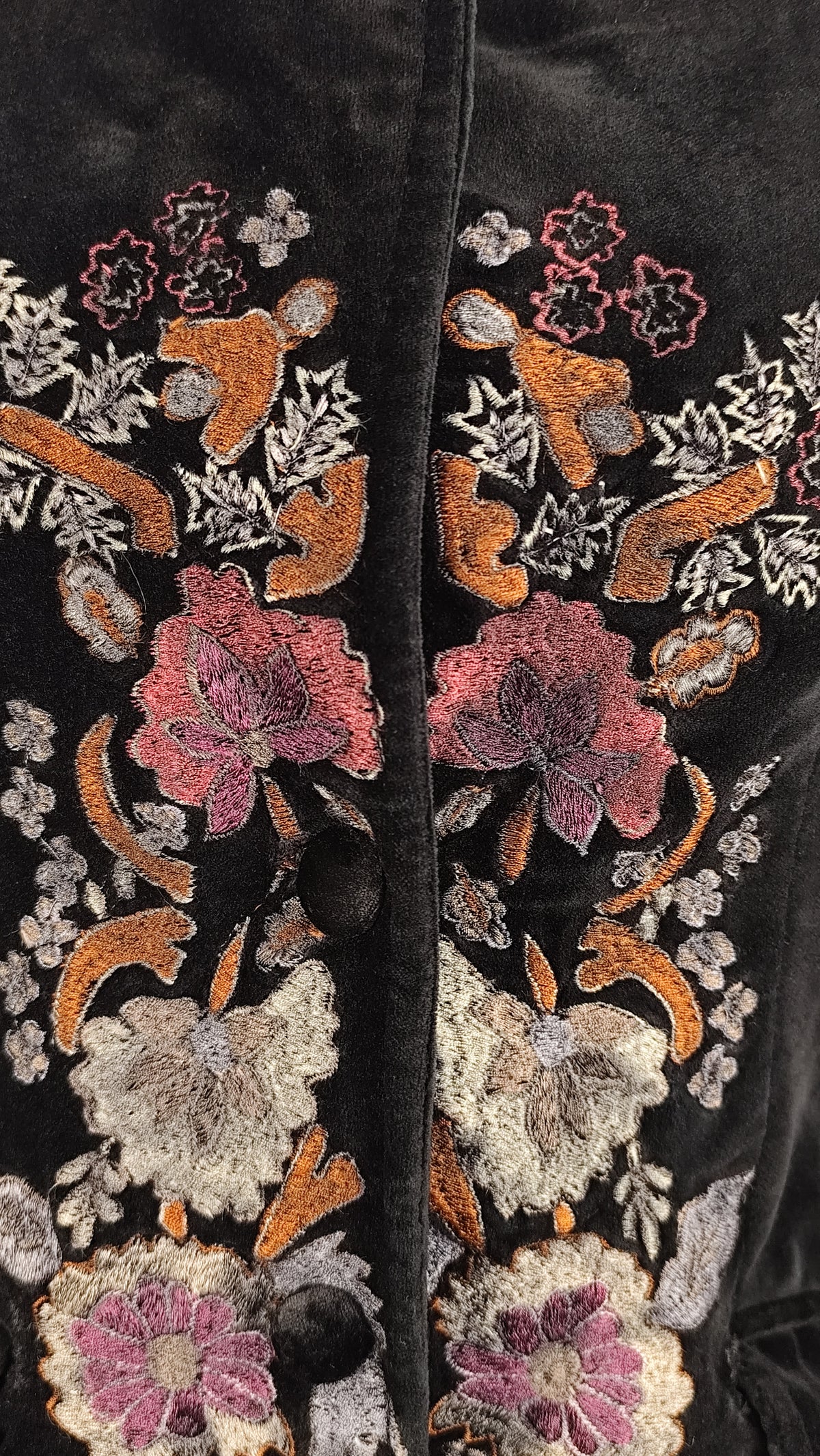 Close-up of a black velvet garment with ornate floral embroidery in pink, orange, beige, and white, featuring a central button closure.