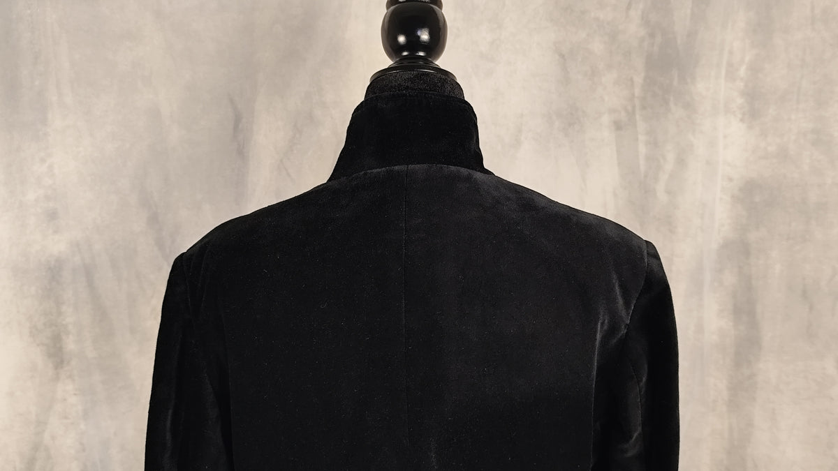 A black velvet coat displayed on a black mannequin torso, viewed from the back against a textured gray background.