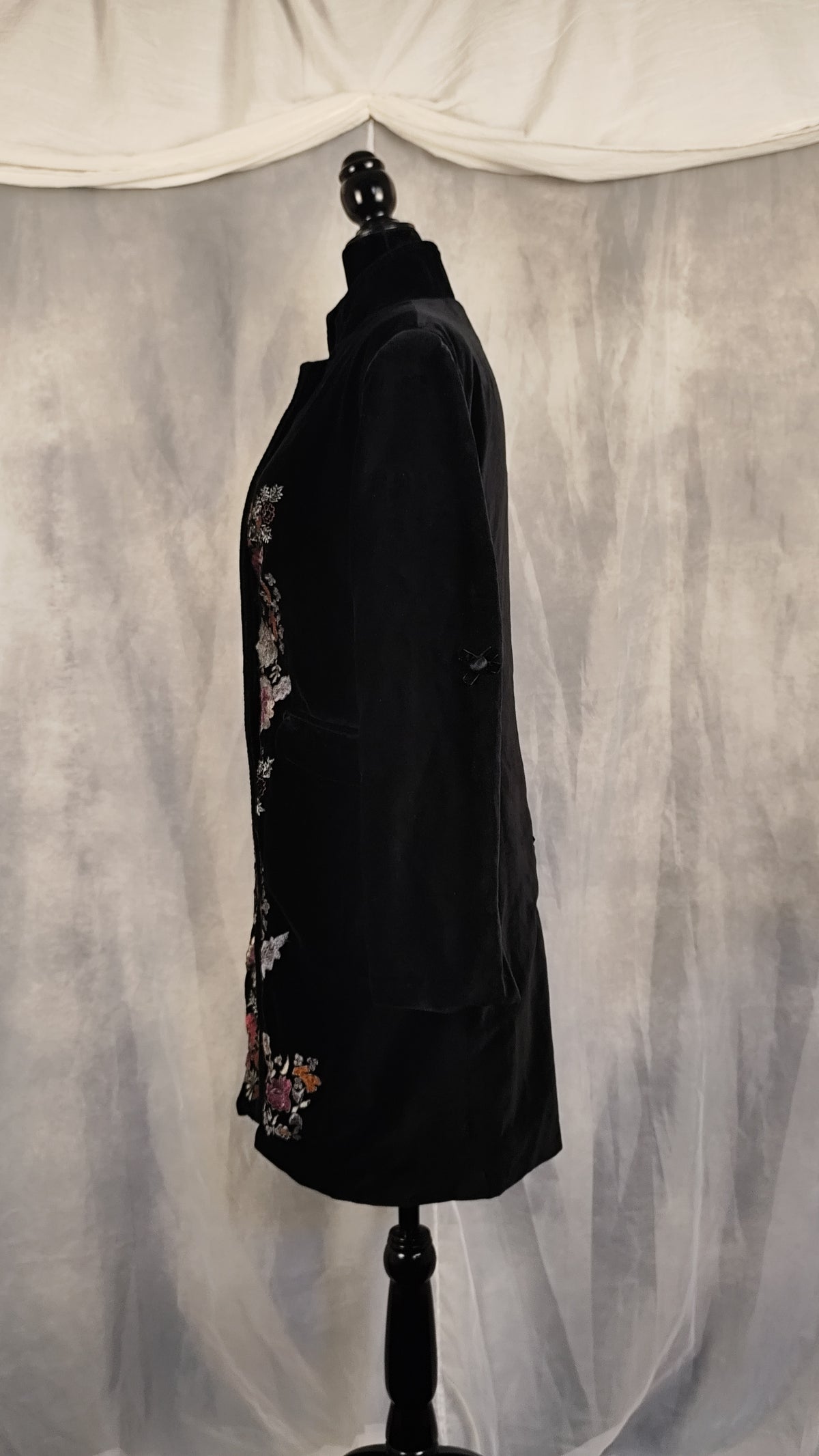 A black longline jacket with colorful floral embroidery on the front and lower hem, displayed on a black dress form mannequin against a gray draped fabric backdrop.